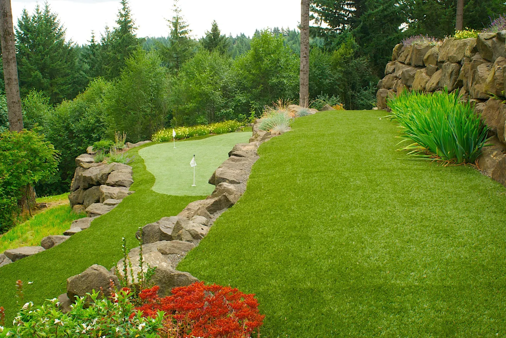 A backyard putting green on a hillside with artificial turf, stone walls, and lush trees.