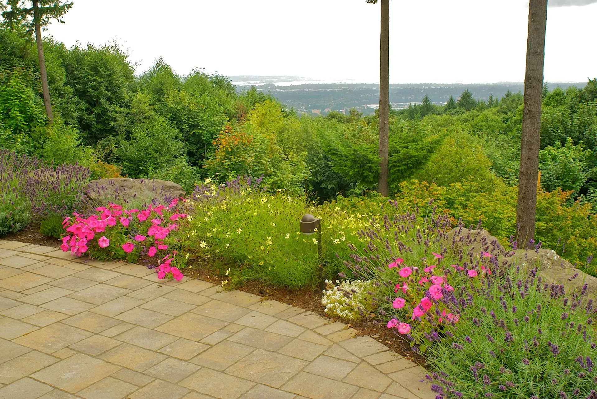 Brick patio overlooking a garden bed of pink and yellow flowers and green foliage, with a distant view of the city.