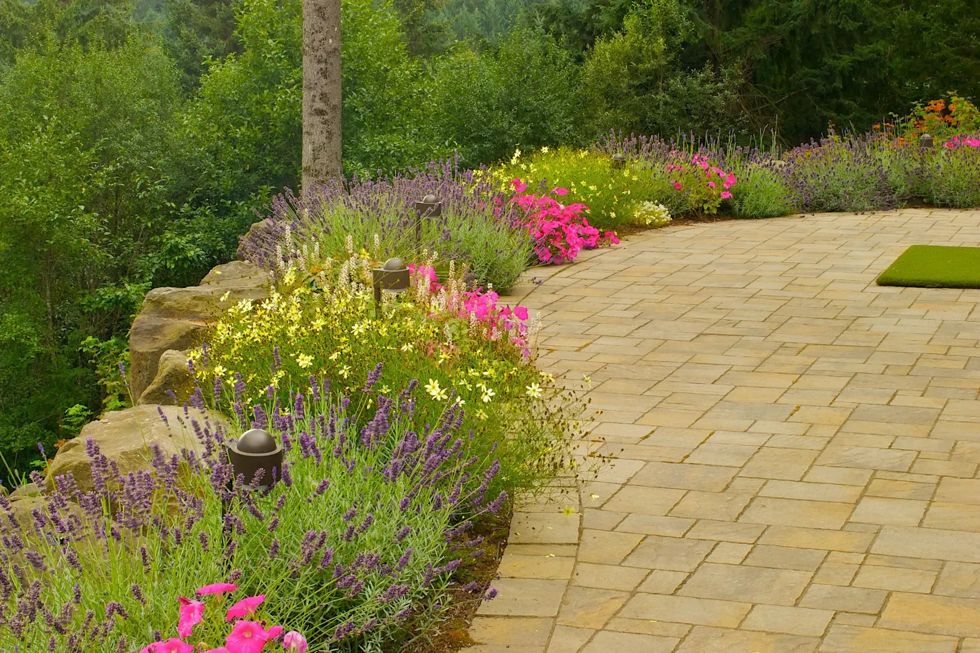 A curved stone patio with colorful flower beds in front of lush greenery and a forest backdrop.