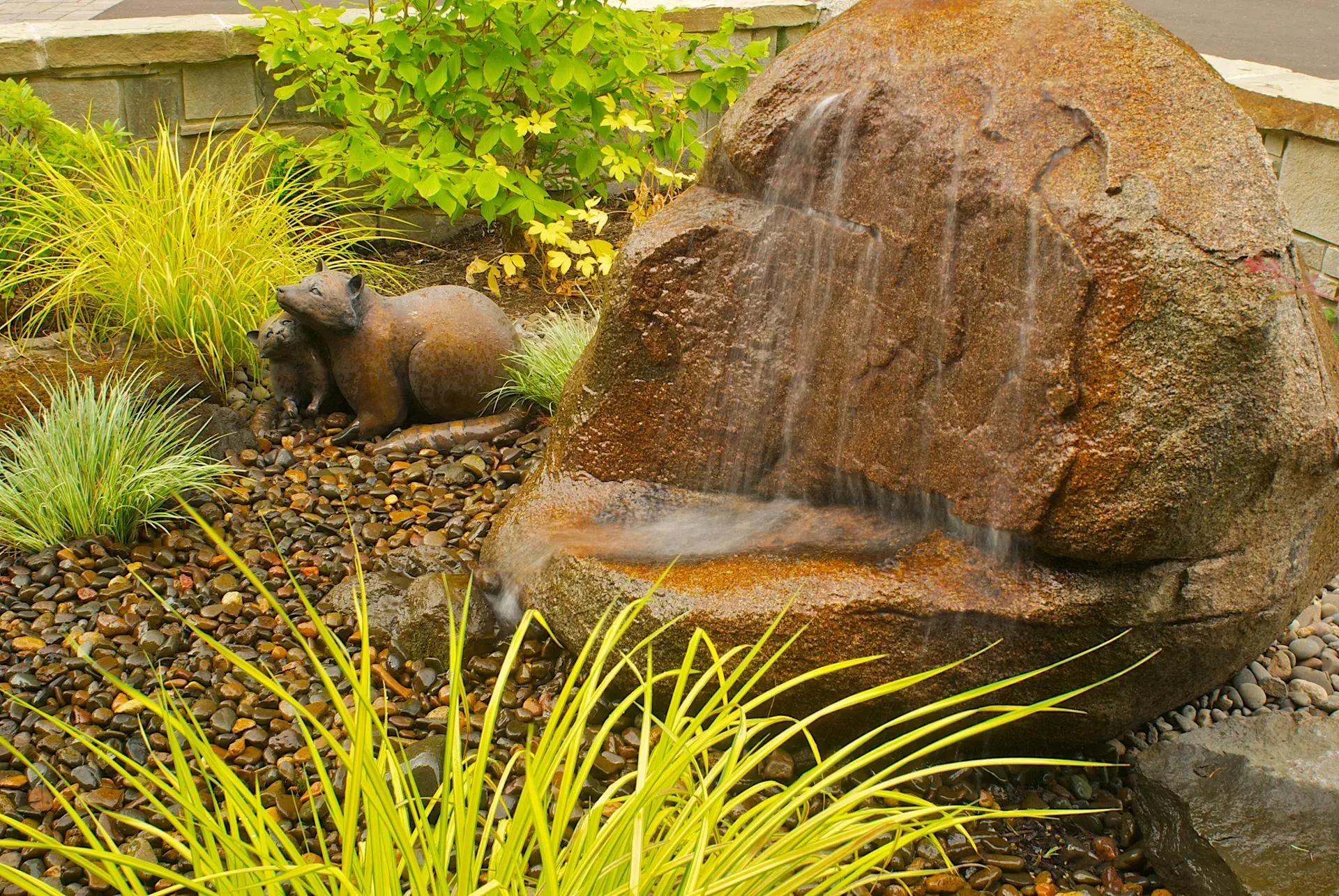 Water fountain feature with a large brown rock, and a brown bear statue among green plants and gravel.