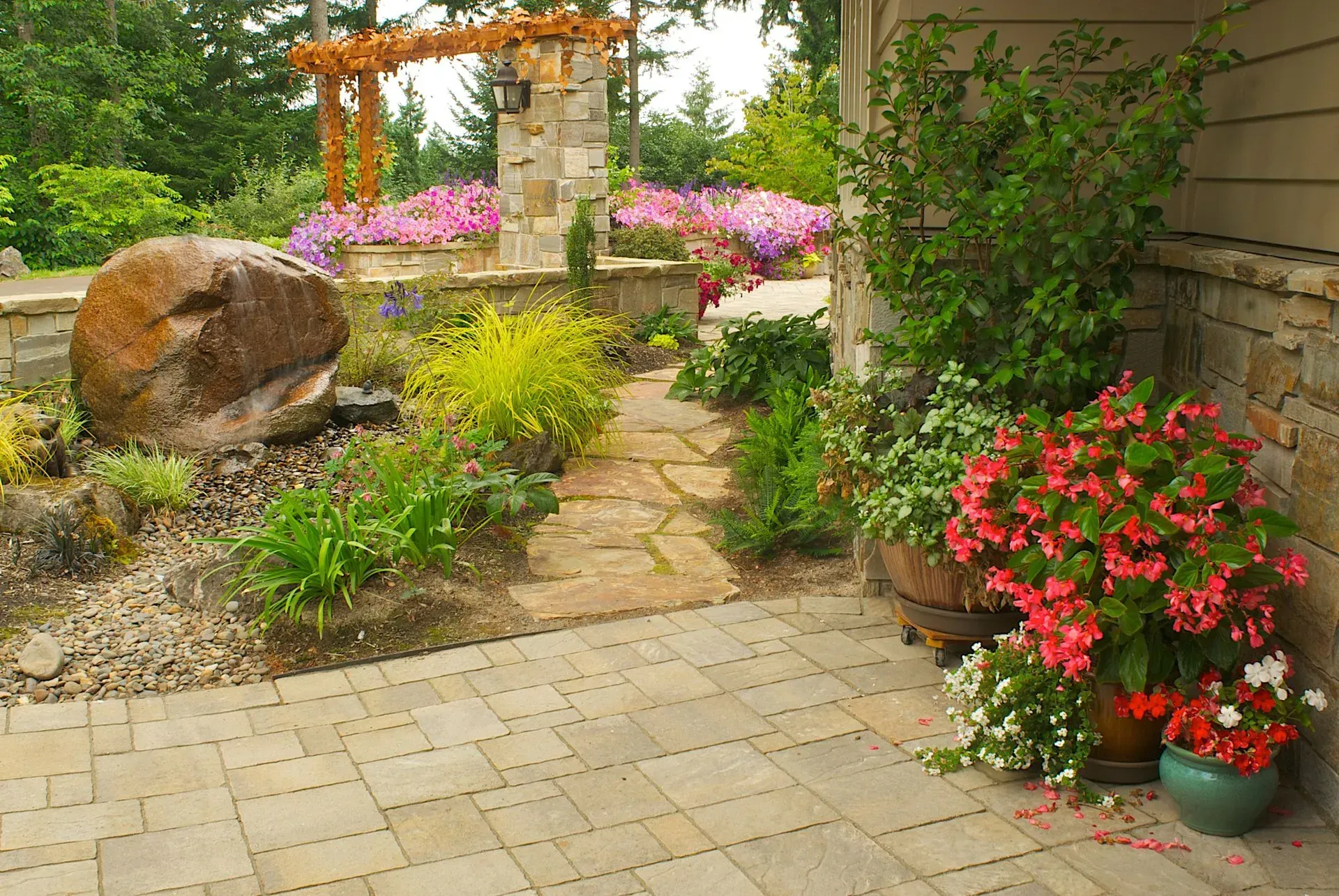 Stone pathway and patio lead to a lush garden with blooming flowers, a boulder, and an arched wooden structure.