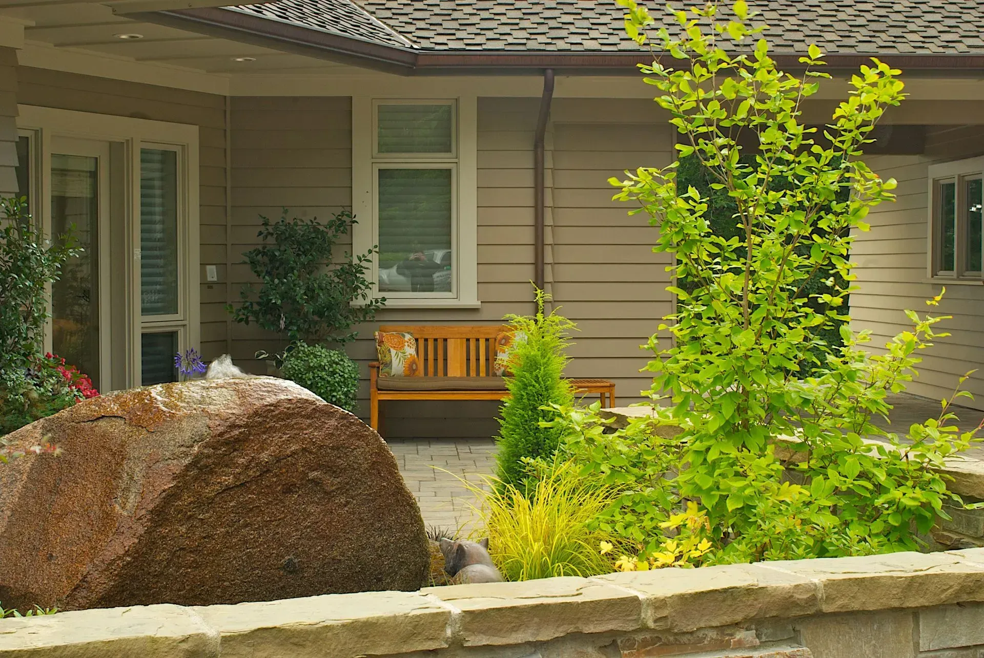 A house exterior with a brown boulder and lush greenery in front of a wooden bench and a window.