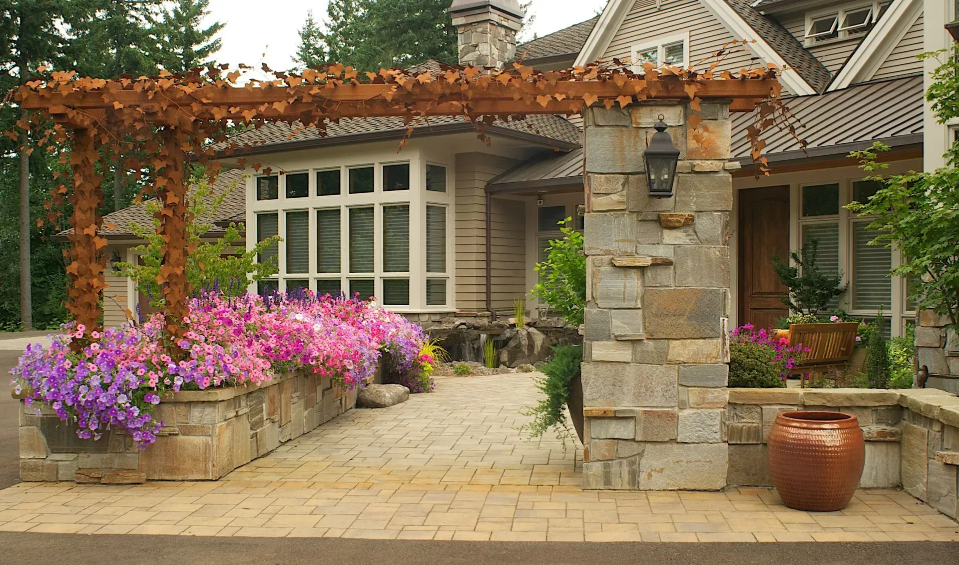 Stone entrance to a house with a pergola covered in vines, surrounded by flowerbeds of pink and purple petunias.