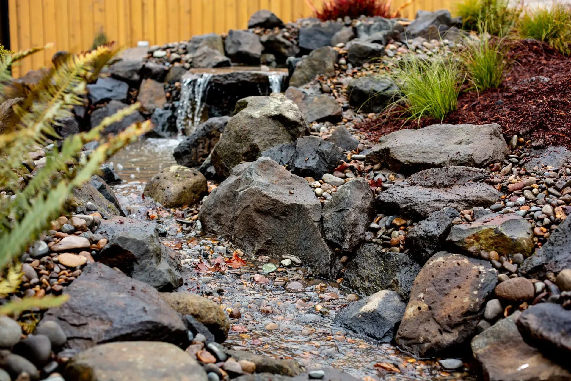 A small waterfall cascades over dark rocks in a garden bed with mulch and plants.
