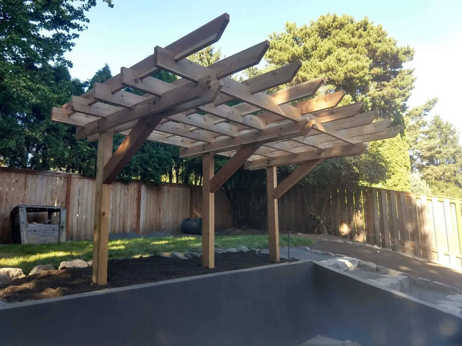 A wooden pergola in a backyard with a wooden fence and trees in the background.