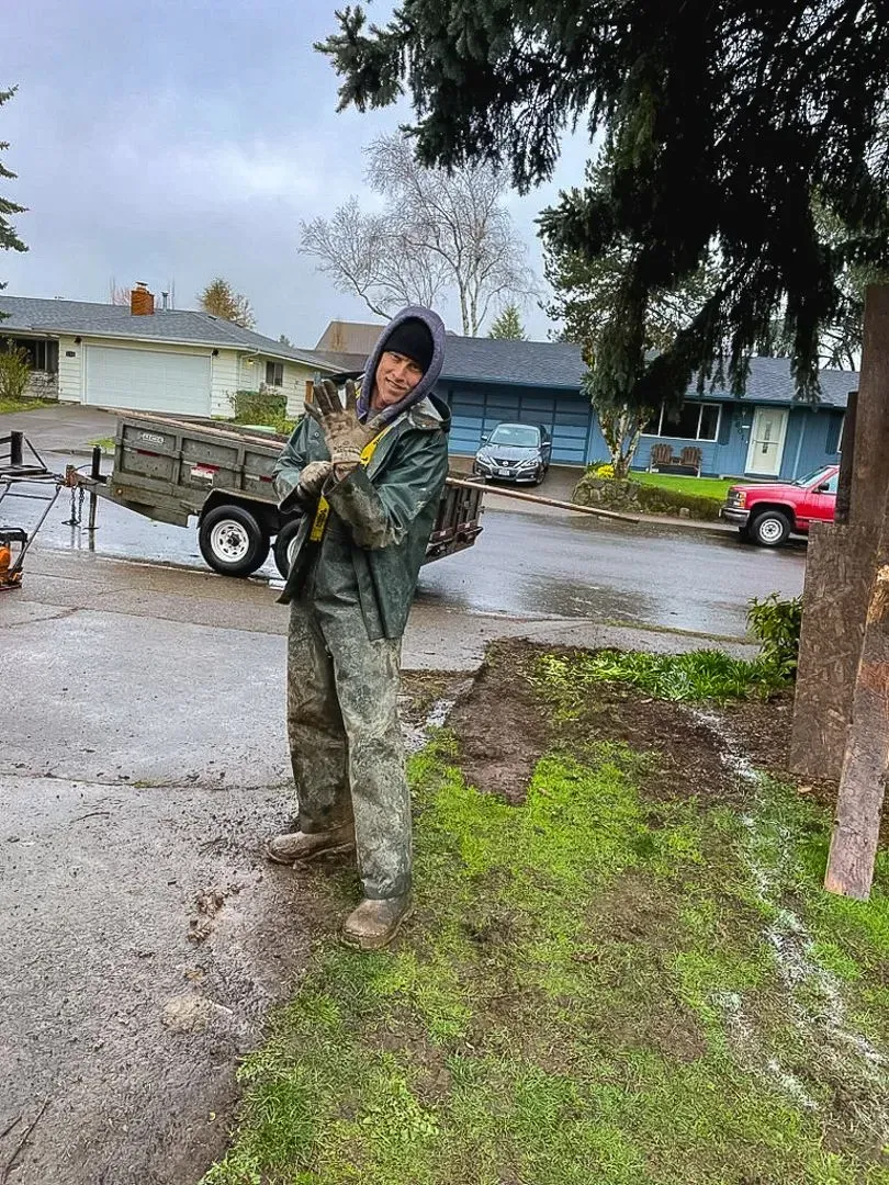 Man in camo pants and jacket smiling, holding a cat outside near a trailer and houses on a cloudy day.