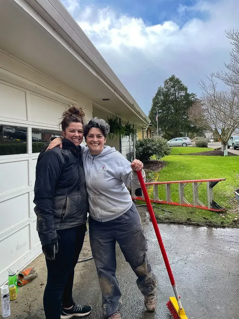 Two women smiling, arm in arm, outdoors. One holds a broom, appearing to have been cleaning. Overcast day.