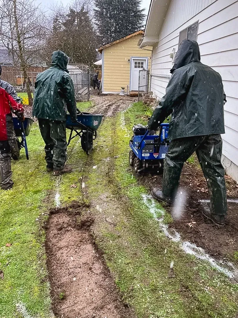 People in rain gear work on a muddy lawn, one using a wheelbarrow, the other a small machine.