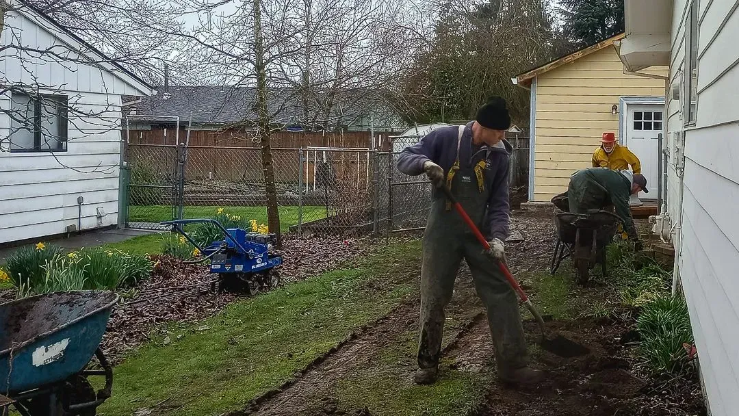Three people working in a yard, one raking dirt, another bending over, and a third standing by a yellow shed.