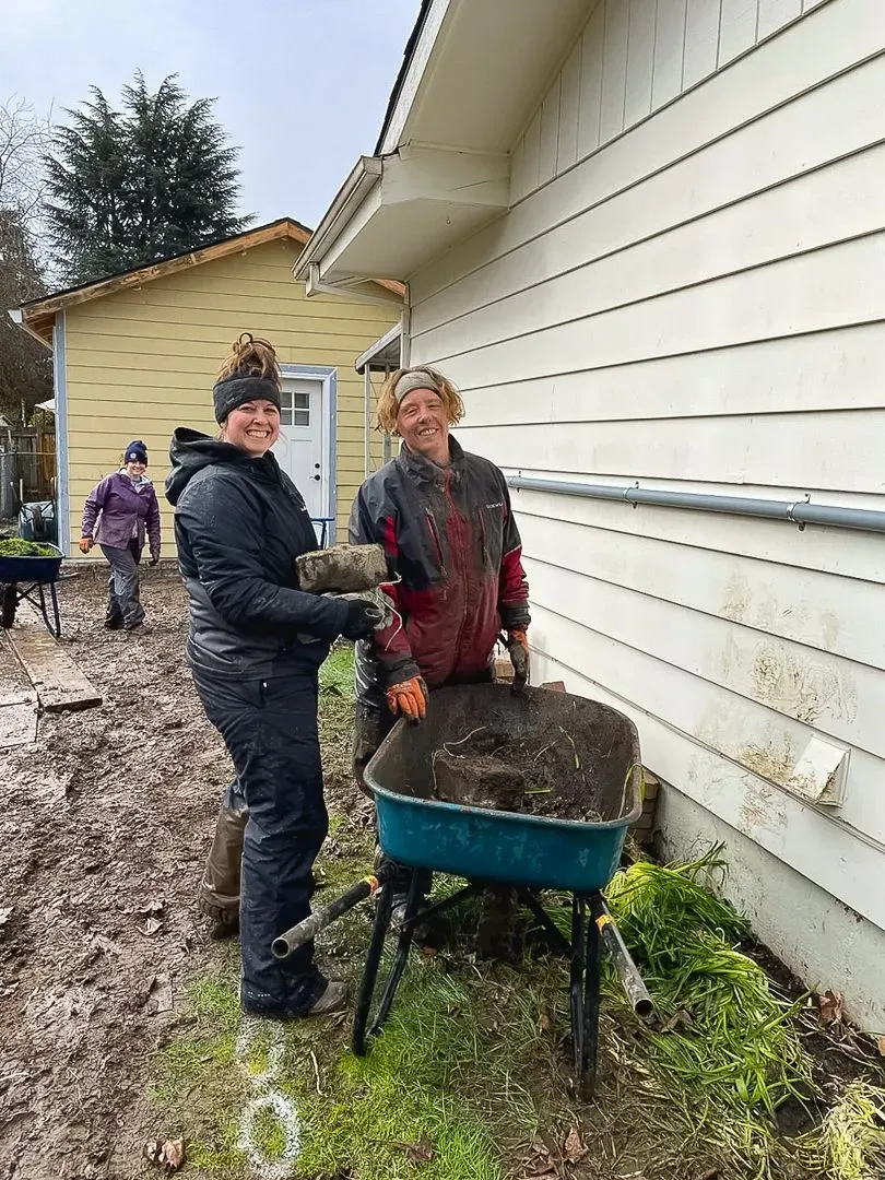 Two people shovel dirt into a wheelbarrow beside a building. The setting is muddy and outdoors on a cloudy day.
