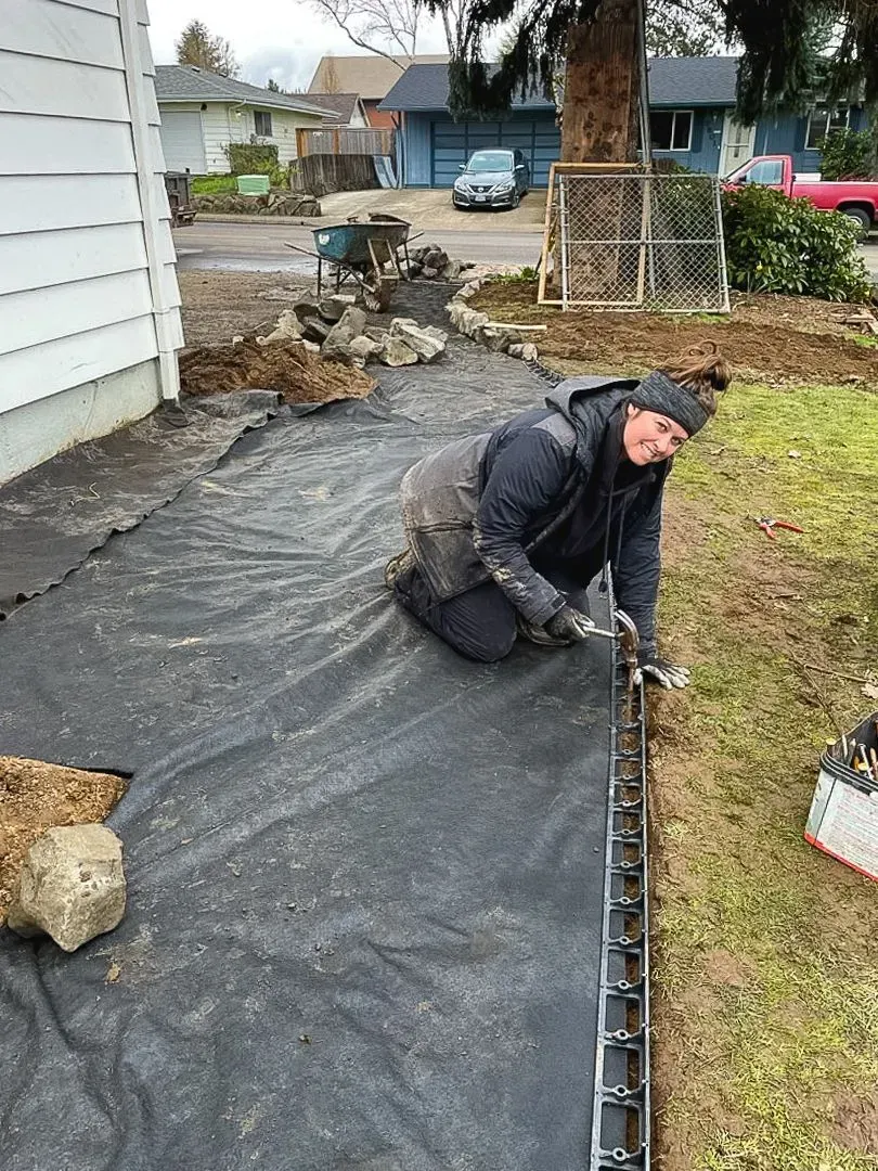 A person kneels on landscaping fabric, smiling while working on a border near a house and lawn.