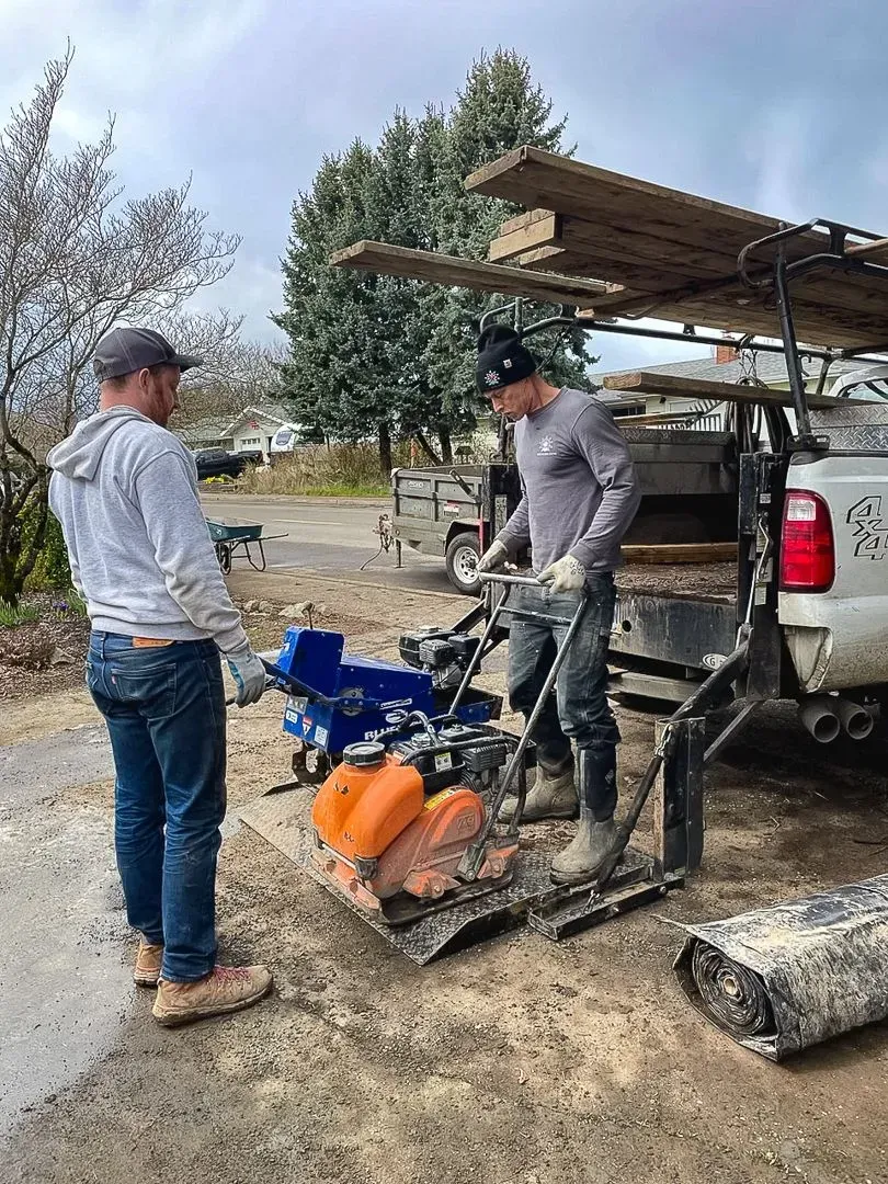 Two men operate a plate compactor near a truck with wood on top.