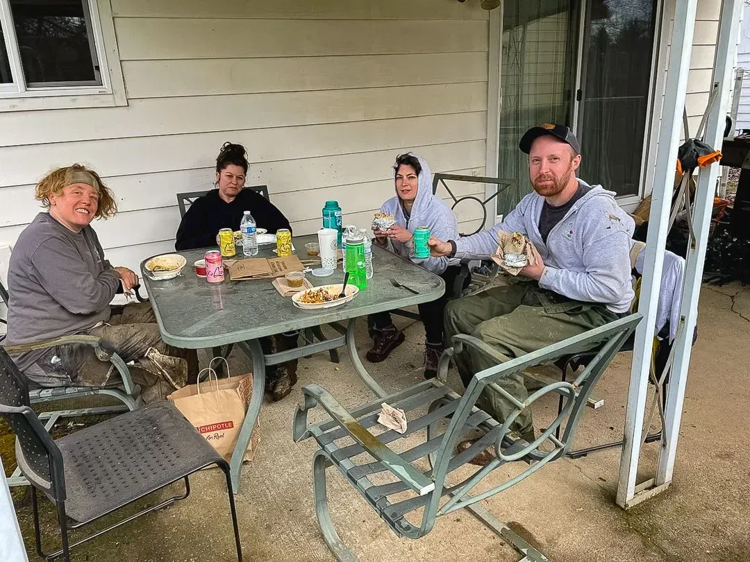 Four people seated around a glass table on a porch eating lunch. They are of varying ages and genders, in casual clothing.