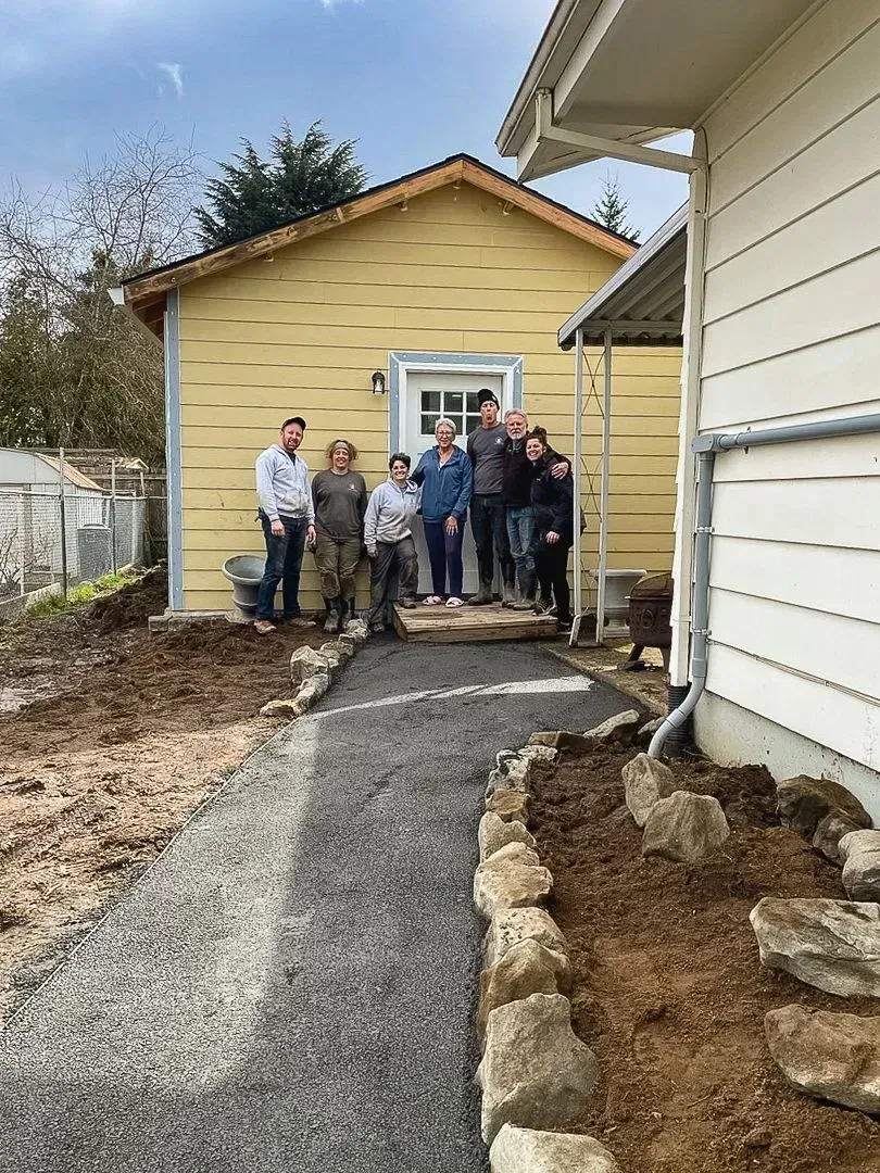 Group of people stand in front of a yellow building, a gravel path leads to the door.