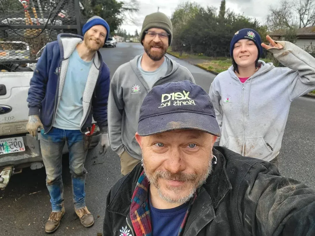 Four people smile for a selfie on a street. The person in the foreground takes the photo, wearing a dirty cap.