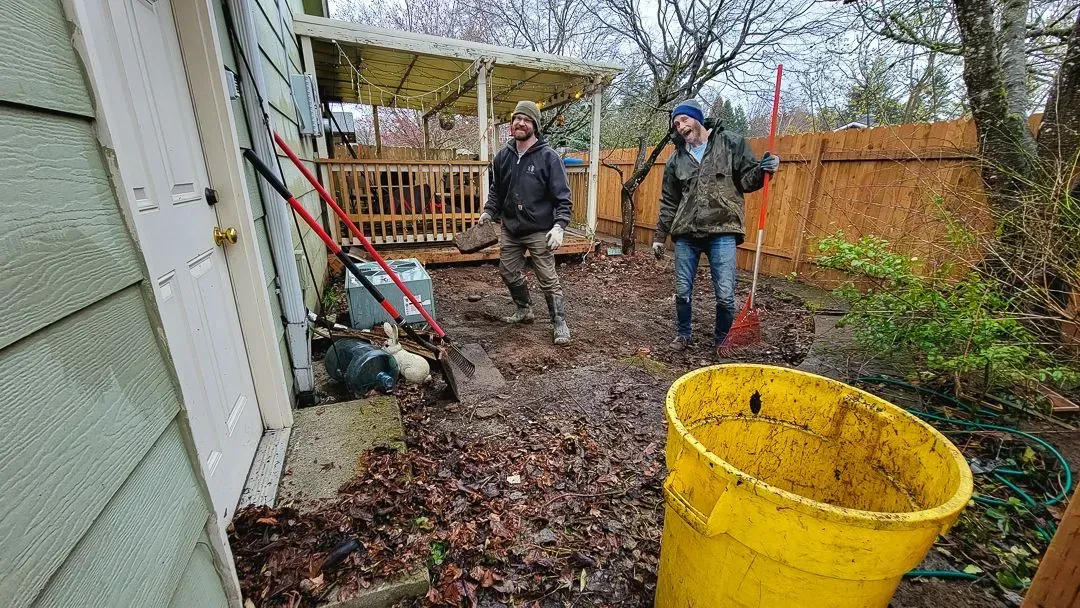 Two people raking leaves in a backyard. A yellow bucket sits near the rakers. The backyard has a wooden fence and a deck.