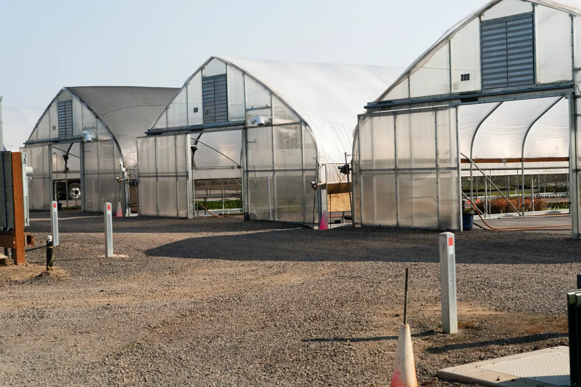 Three connected greenhouses with arched roofs stand on a gravel lot under a clear sky.