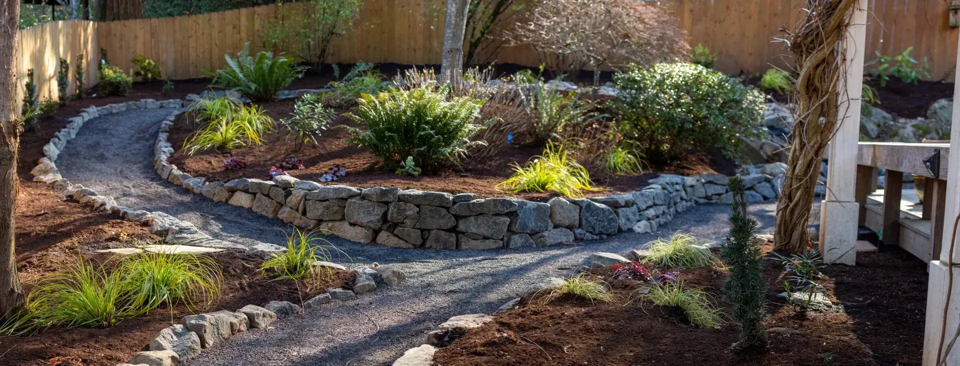 A winding gravel path in a garden, flanked by flower beds and a stone retaining wall. 