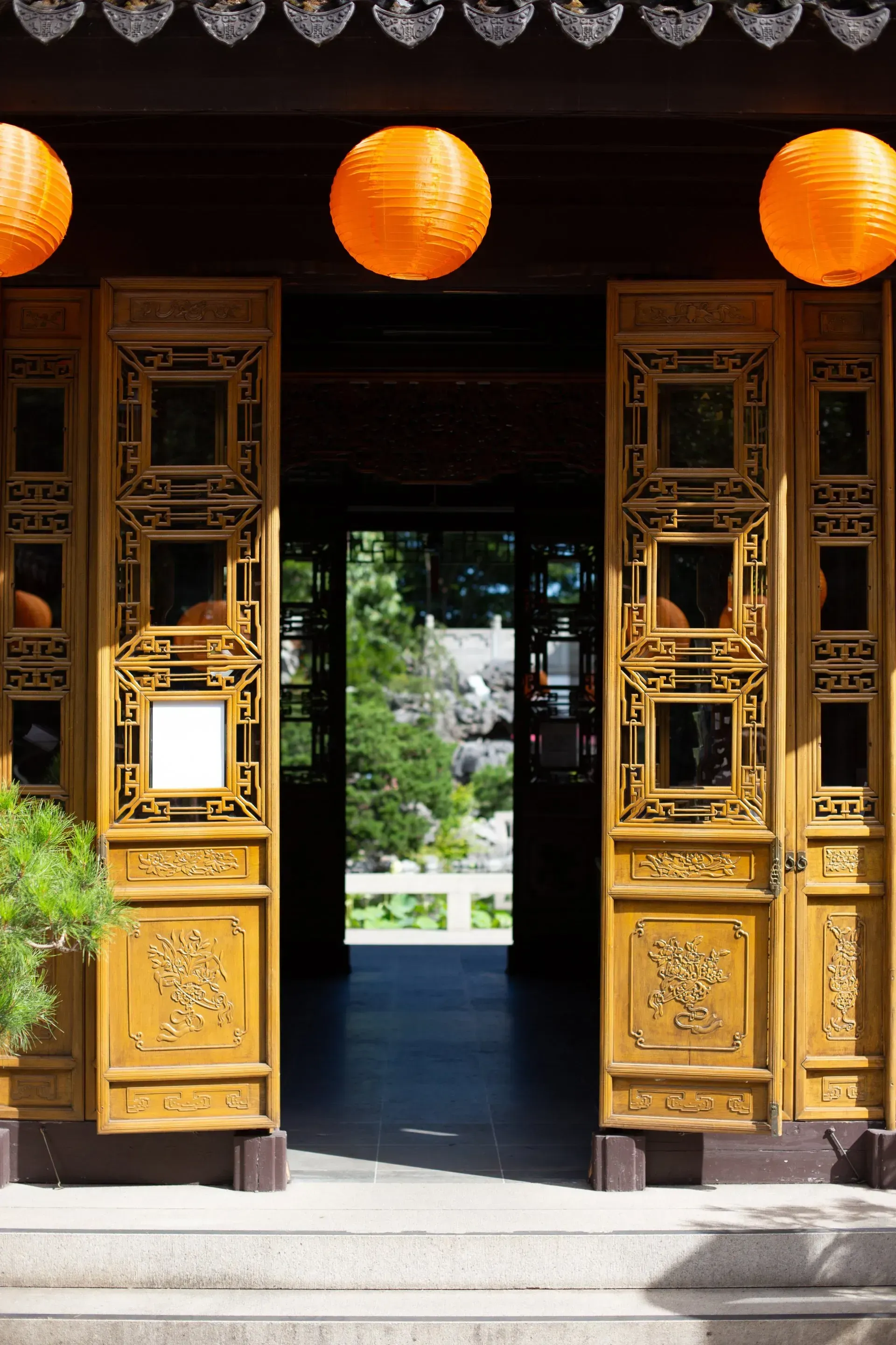 Ornate wooden doors of a Chinese-style building, orange lanterns hanging above the entrance, leading to a garden view.