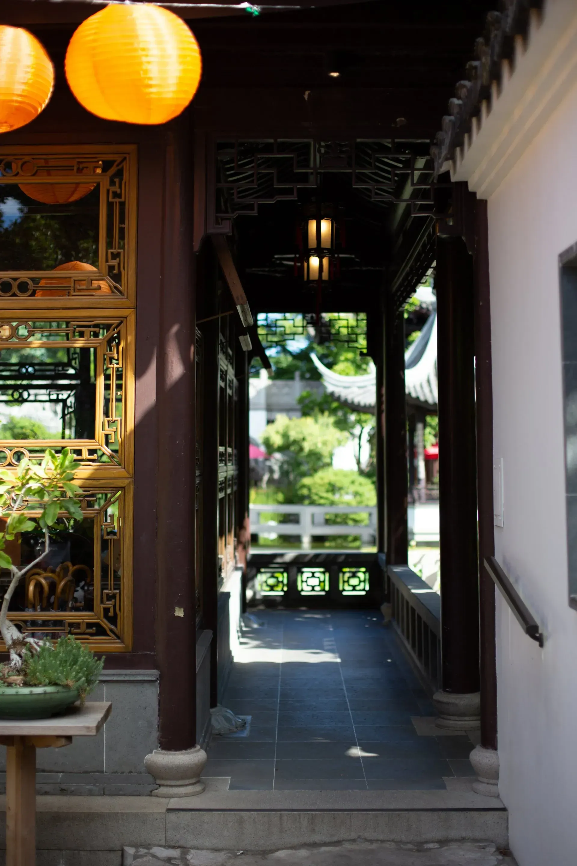 A narrow, shaded corridor in a traditional Asian garden, with decorative lanterns and a view of a garden and bridge.
