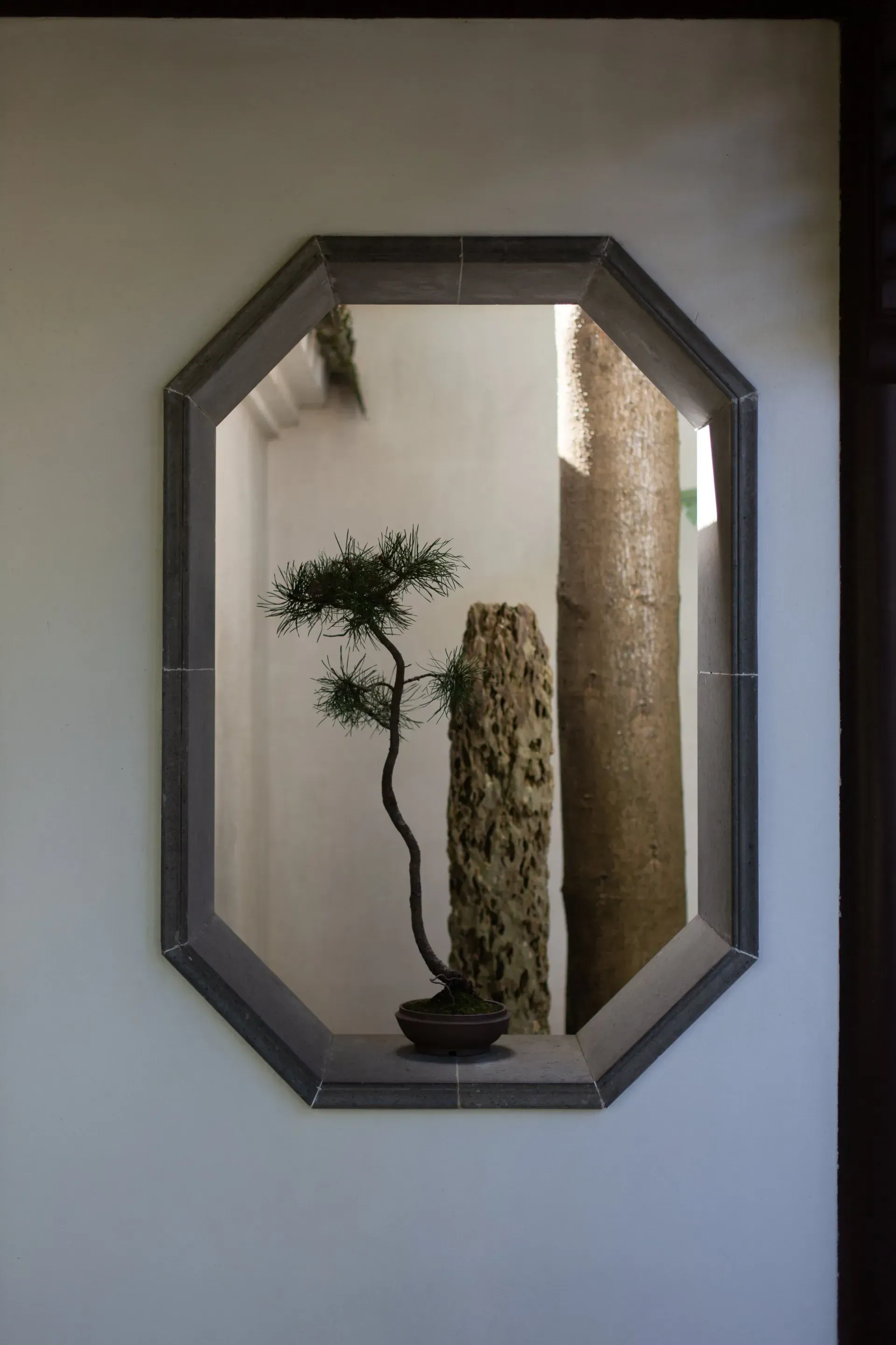 An octagonal window frame on a white wall displays a bonsai tree and a carved stone, inside a courtyard.