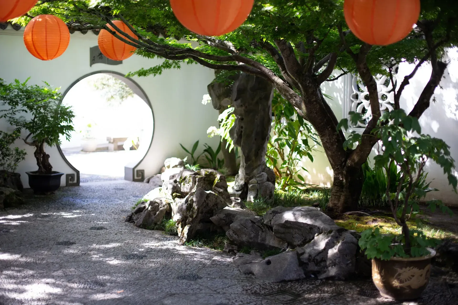 A serene garden with a round doorway, orange lanterns, and a stone pathway beneath a tree's canopy.