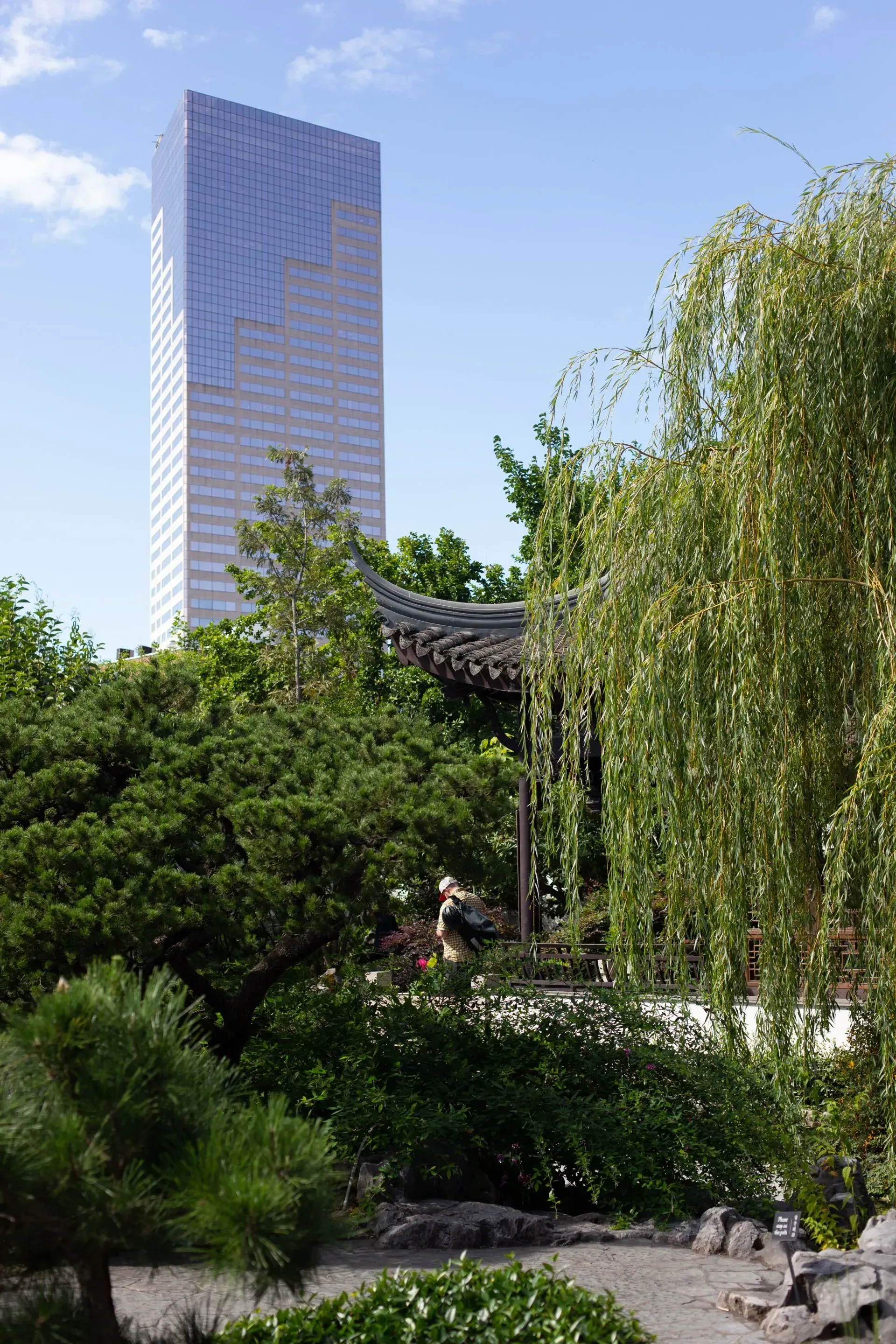 Tall glass skyscraper rises above a lush garden with trees and a decorative structure; blue sky in the background.