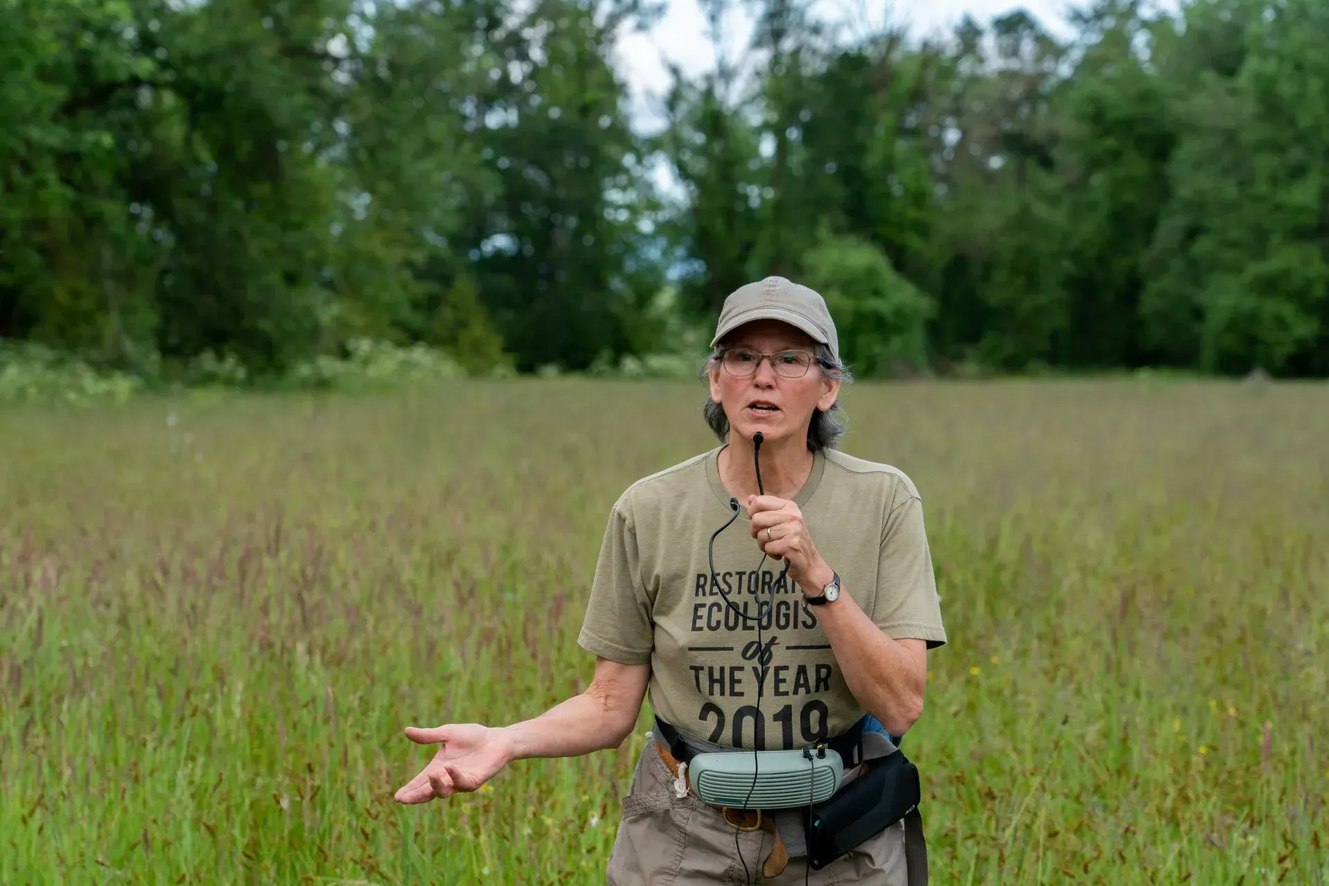Woman in cap and t-shirt speaks into a mic in a grassy field, gesturing with hands. Trees are in the background.