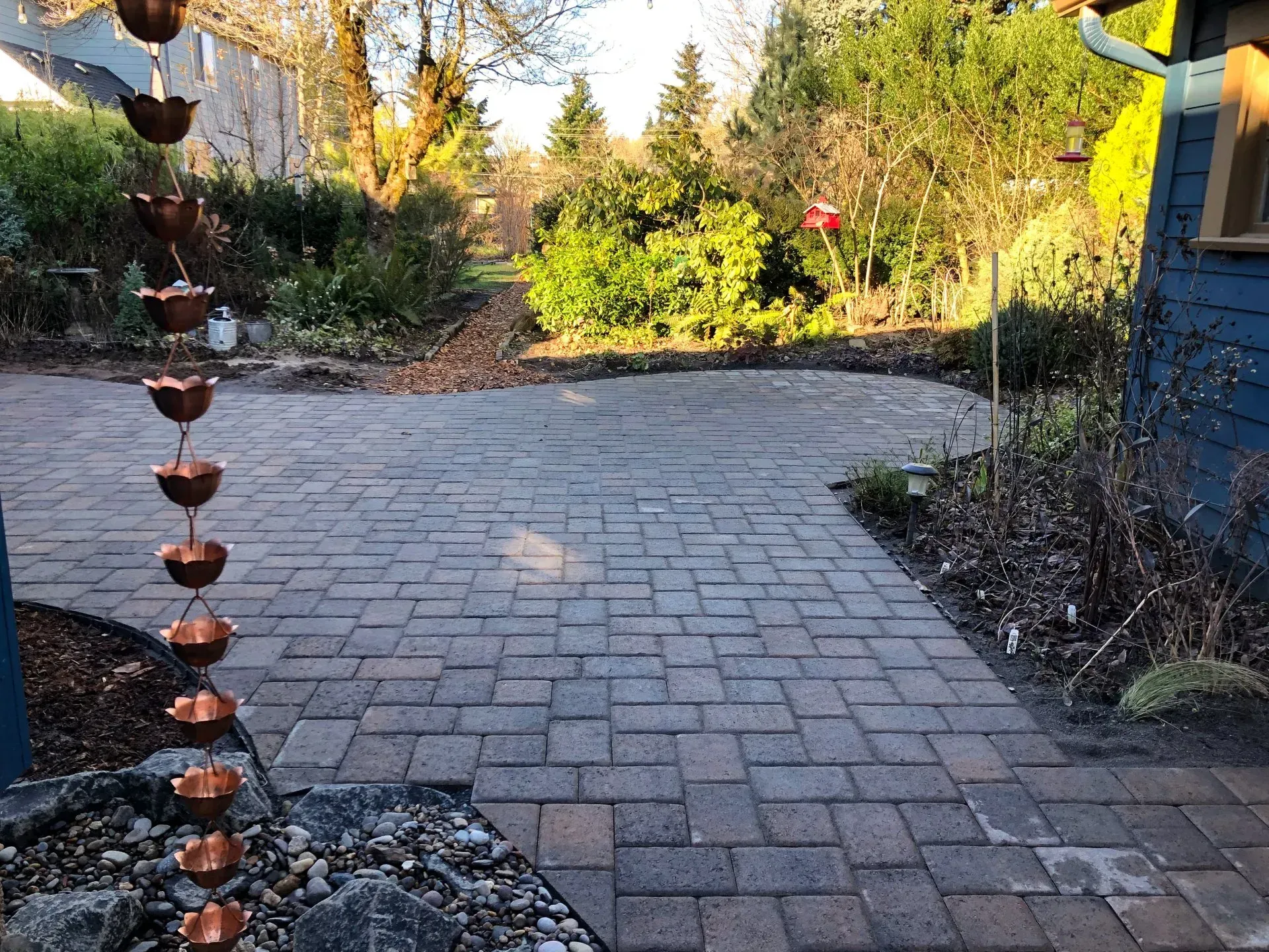 Brick patio with copper rain chain, leading to a garden with trees and a blue building.