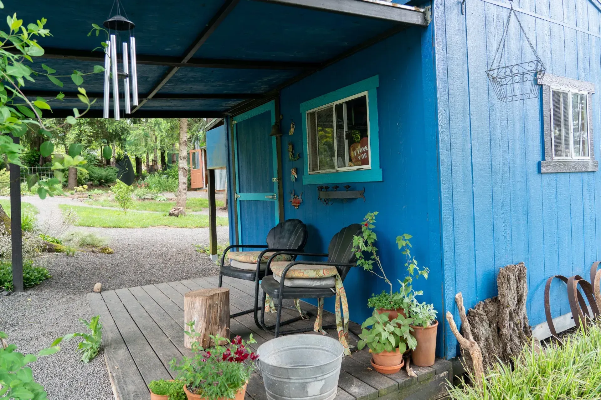 Bright blue cabin with a covered porch, two chairs, potted plants, and a view of a green yard. 