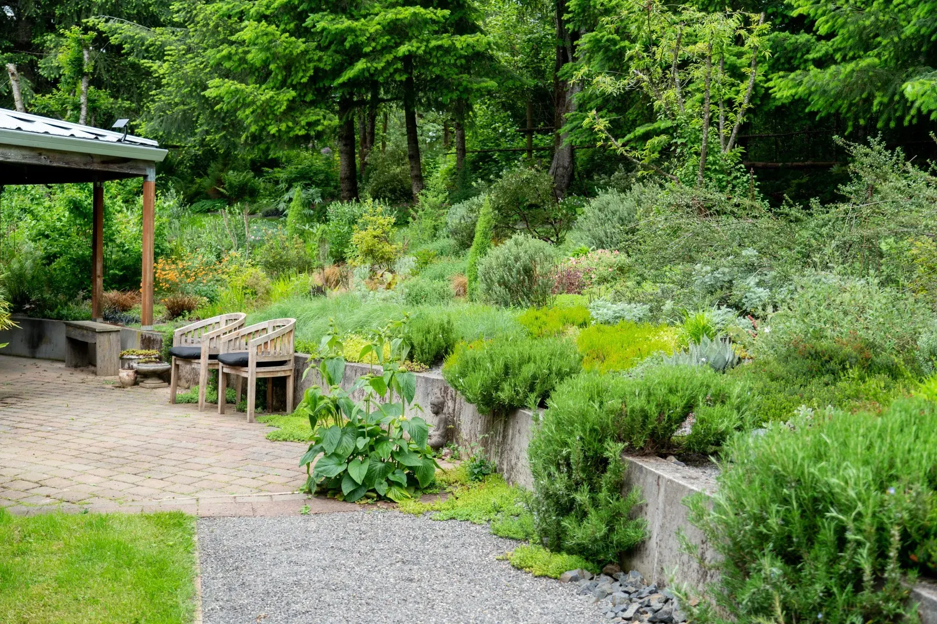 A terraced garden with lush greenery, benches, and a gravel pathway next to a lawn; trees in the background.