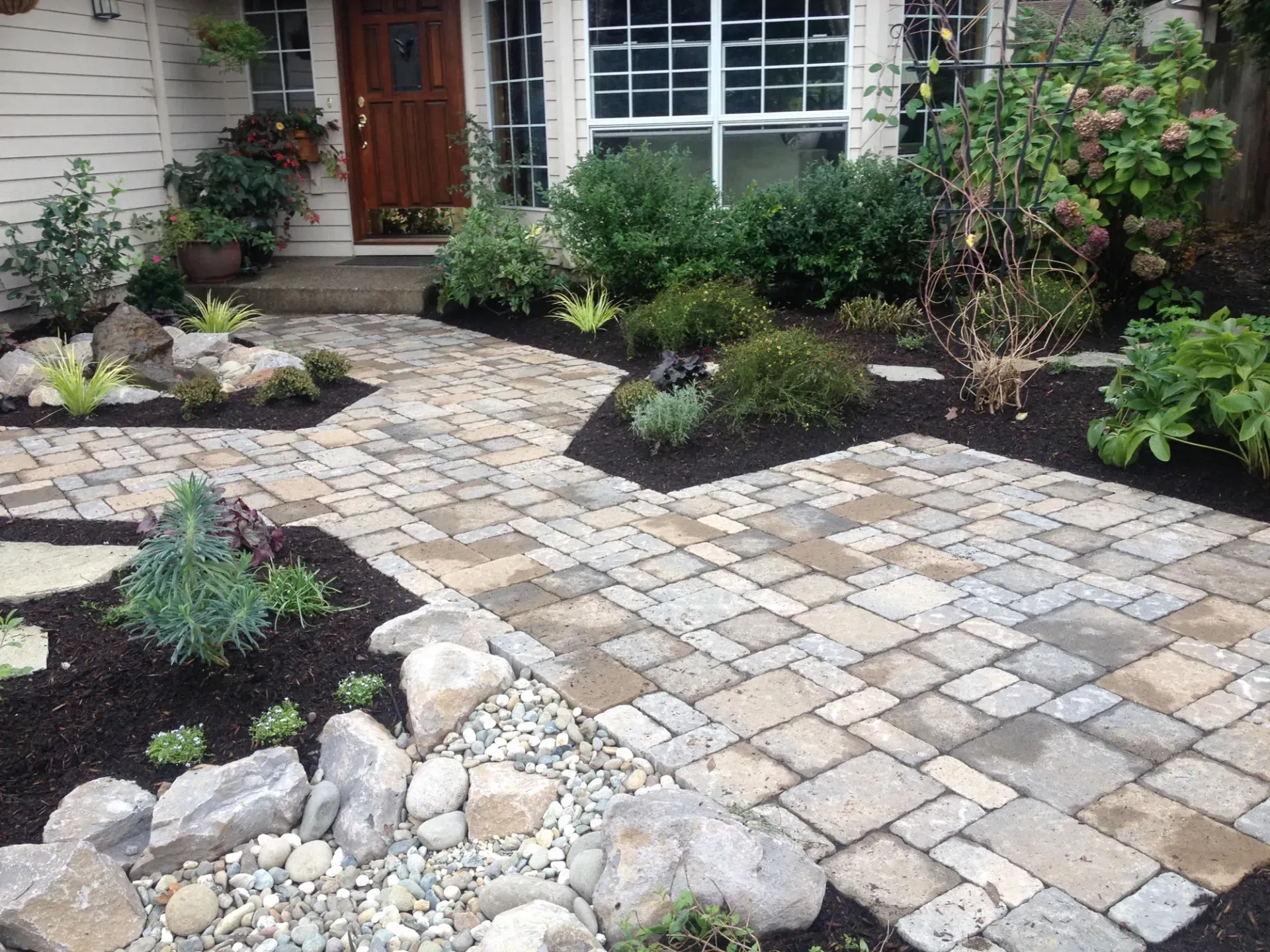 Brick walkway leading to a house entrance surrounded by landscaped beds with rocks, mulch, and greenery.