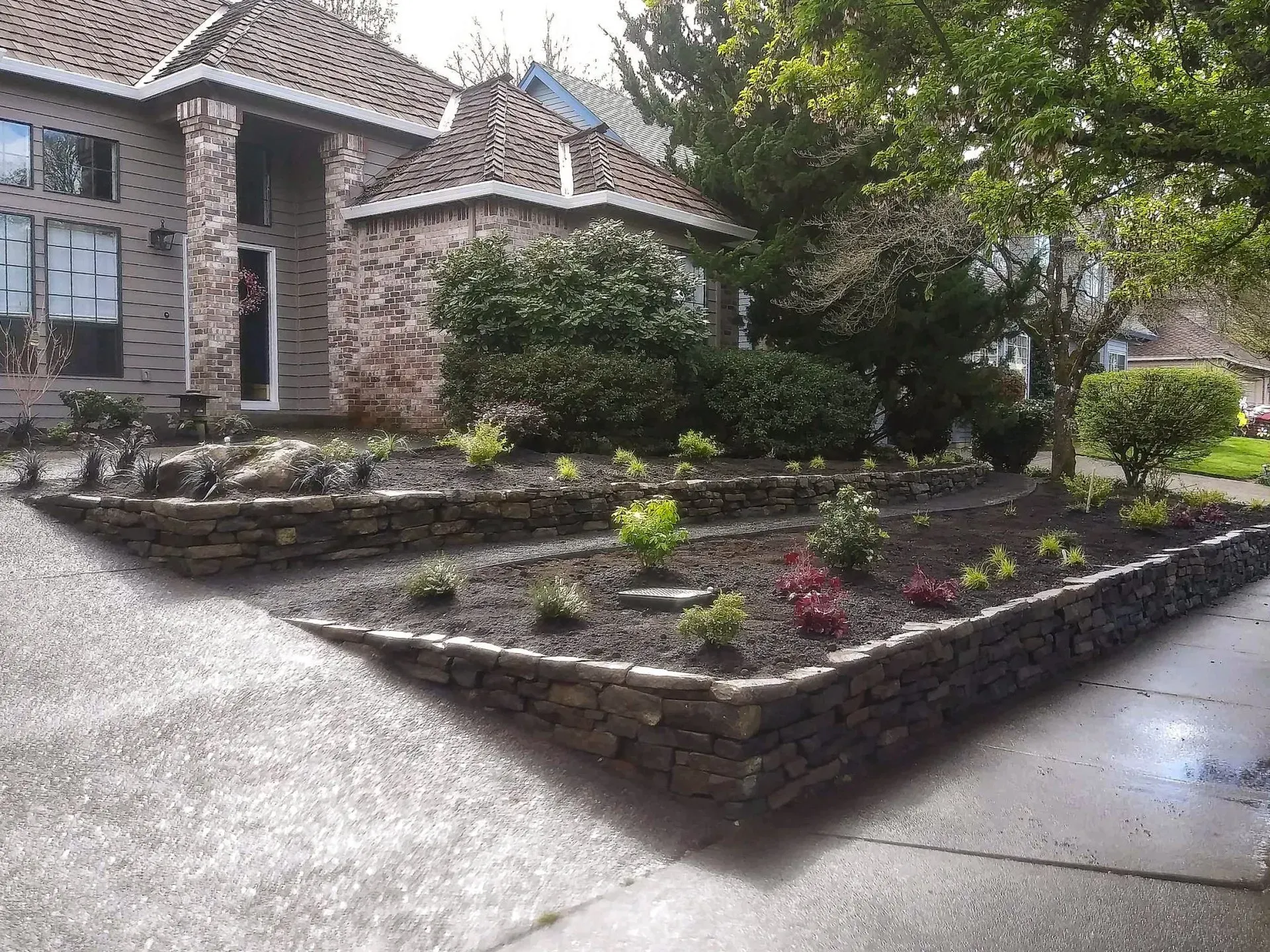 Stone-edged flower beds in front of a brick house with a gray driveway.
