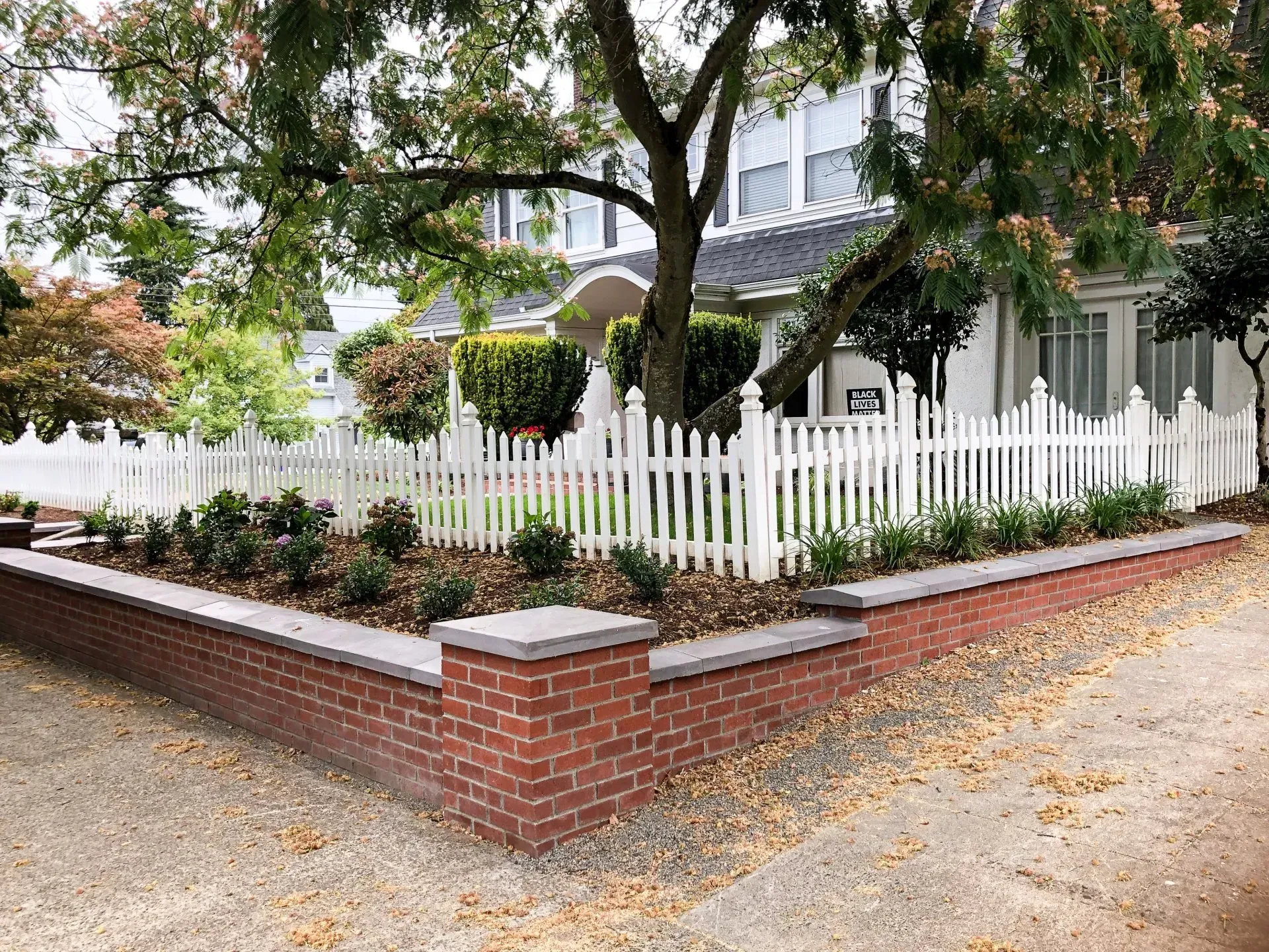 Brick and stone front yard with a white picket fence and a house in the background.