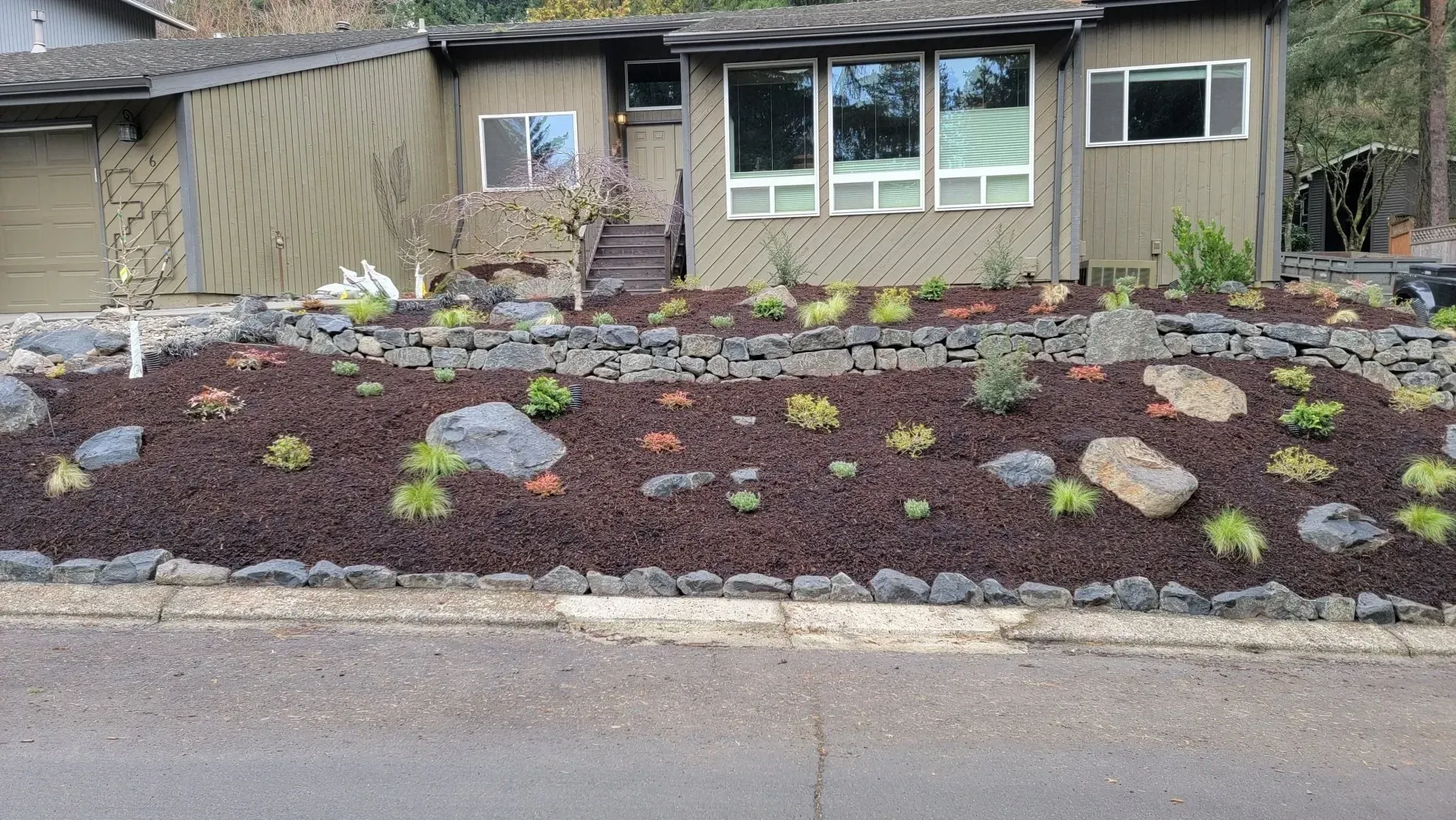 A front yard with a rock retaining wall, mulch, and plants in front of a brown house. Gray rocks are scattered throughout.