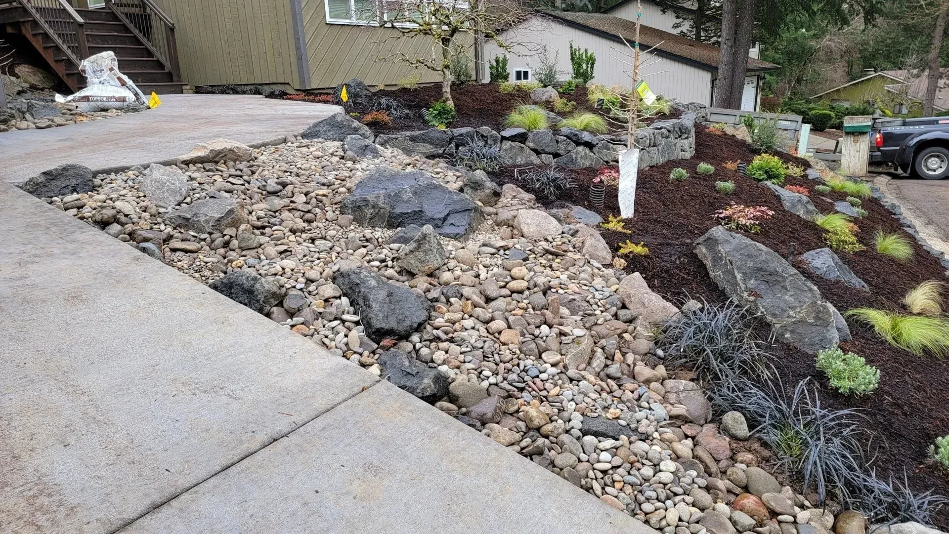 A driveway leads to a rock and mulch landscaped garden with plants and large boulders.