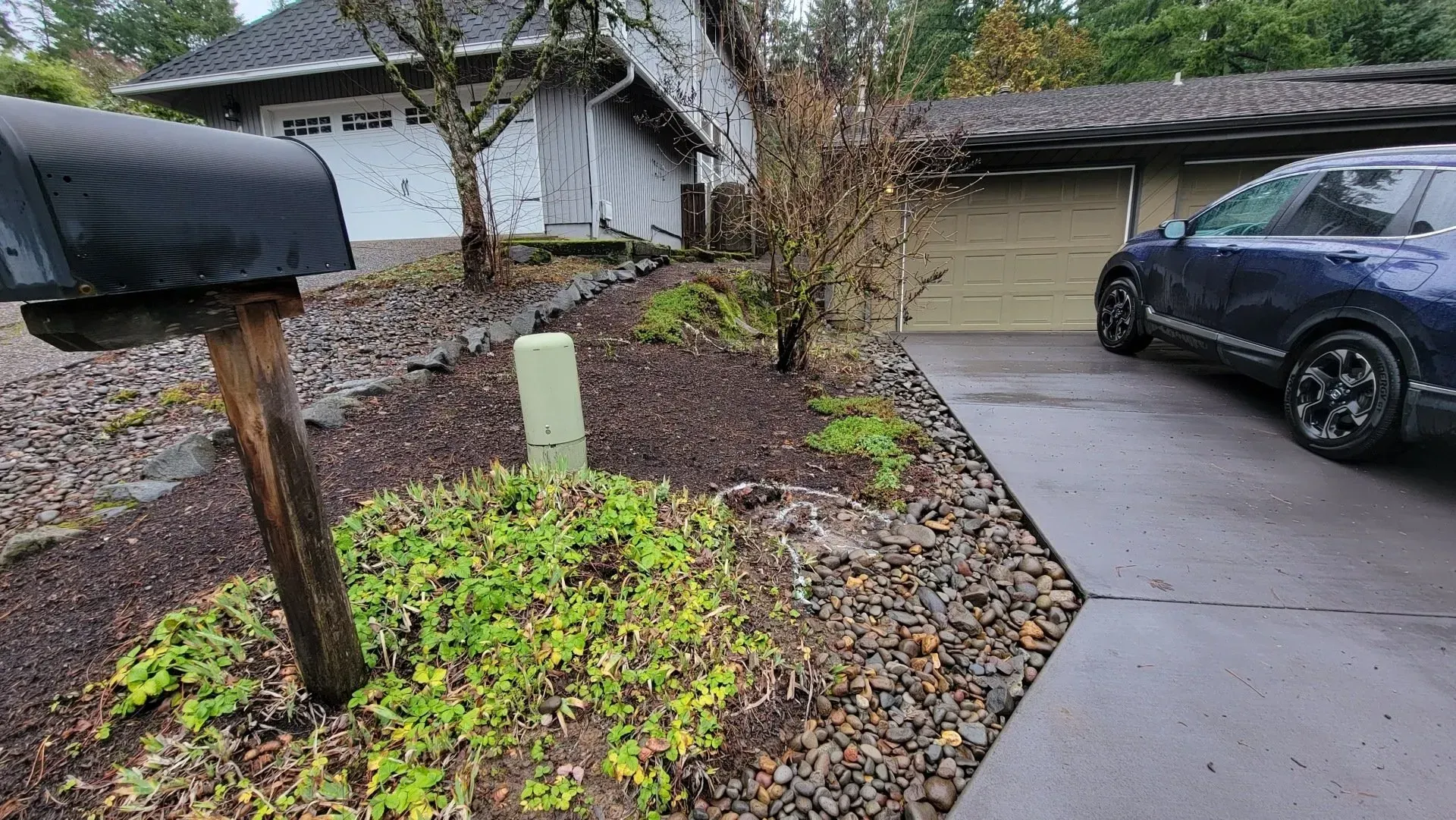 A blue car parked in a driveway next to a house with a brown yard and mailbox in the foreground.