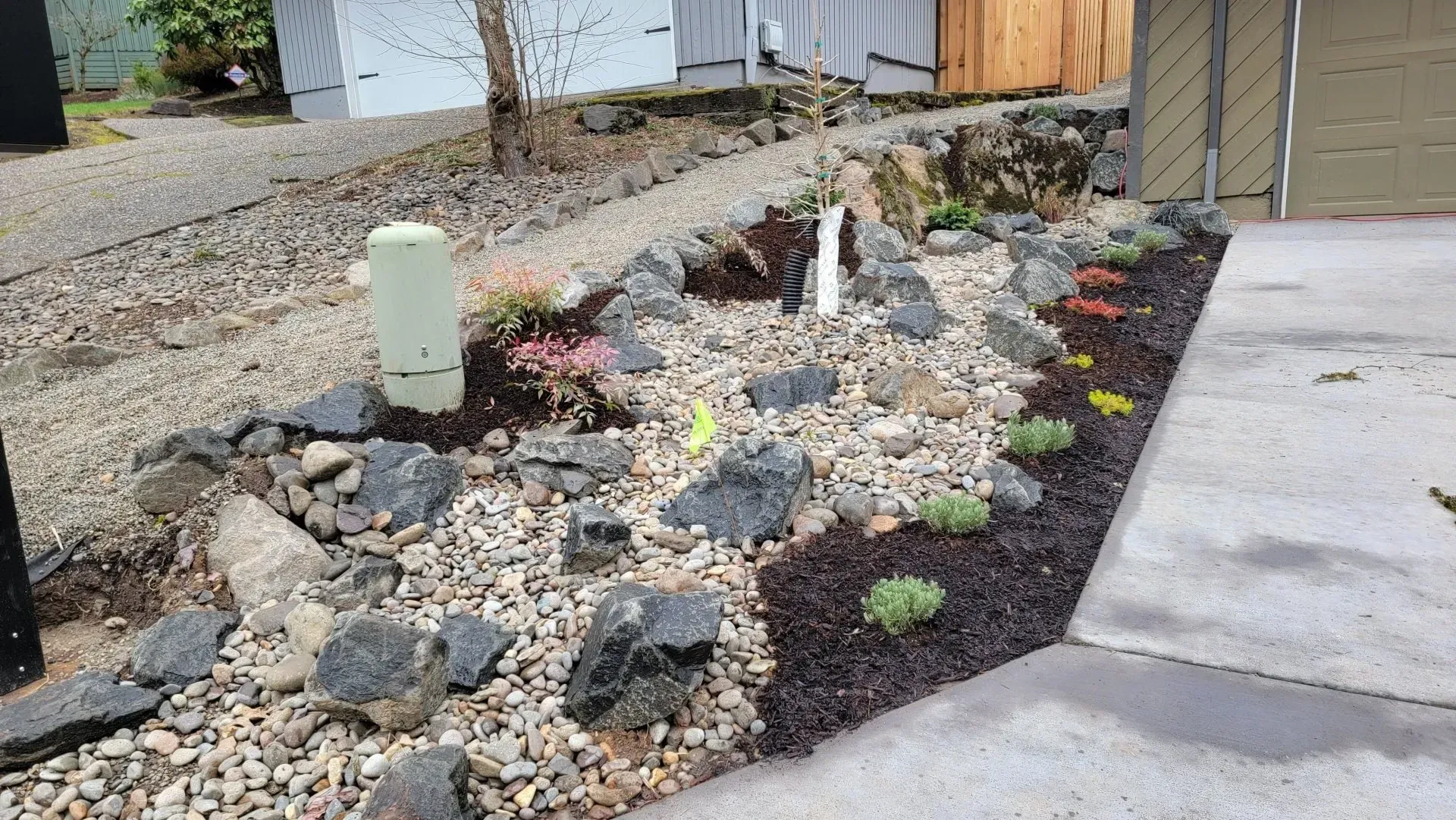 A rock garden with small plants and mulch next to a driveway and road. An electrical box is in the garden.