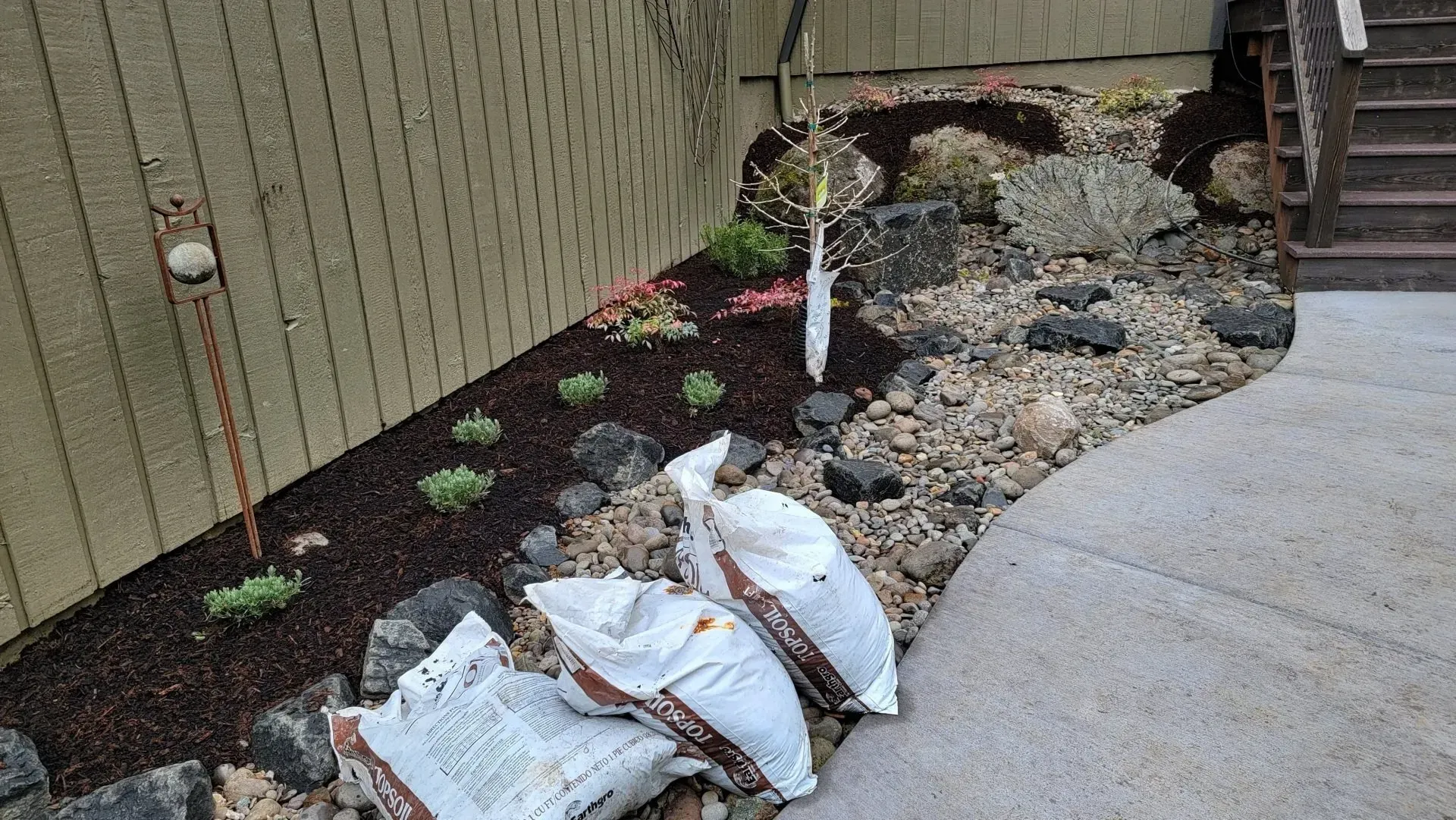 Garden bed with rocks, mulch, and small plants next to a stucco wall and concrete path; three bags of soil sit in front.