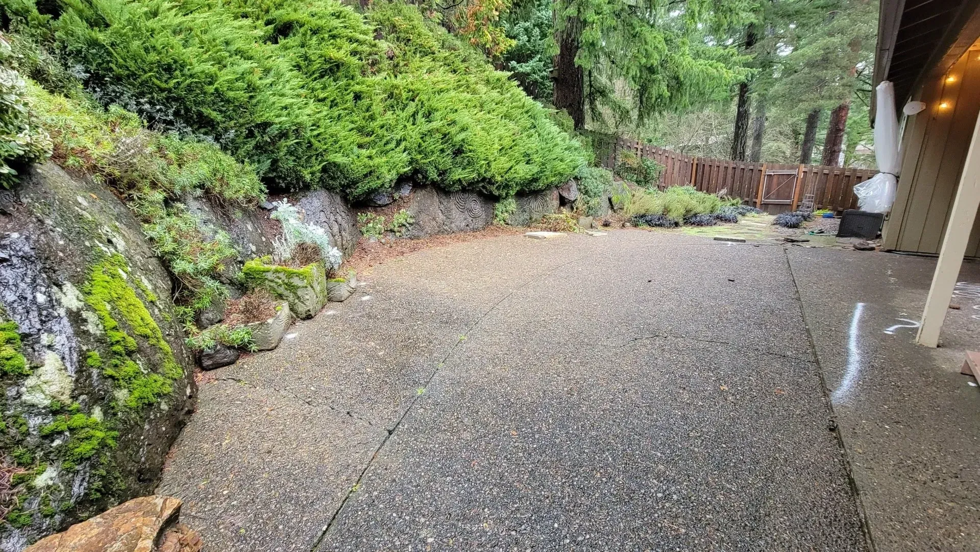 A gravel patio borders a mossy rock wall with greenery, leading to a wooden fence in a wooded setting.