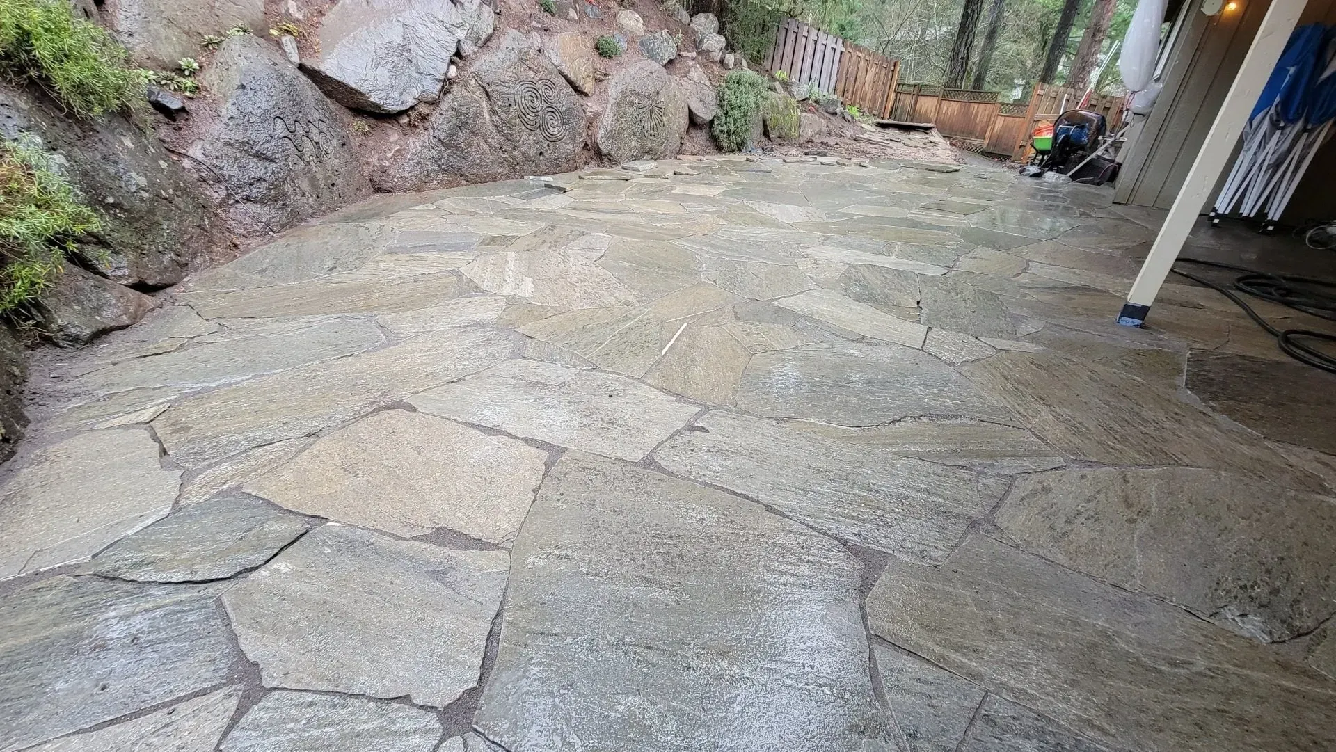 Stone patio with uneven flagstones next to a mossy rock wall, with a wooden deck and trees in the background.
