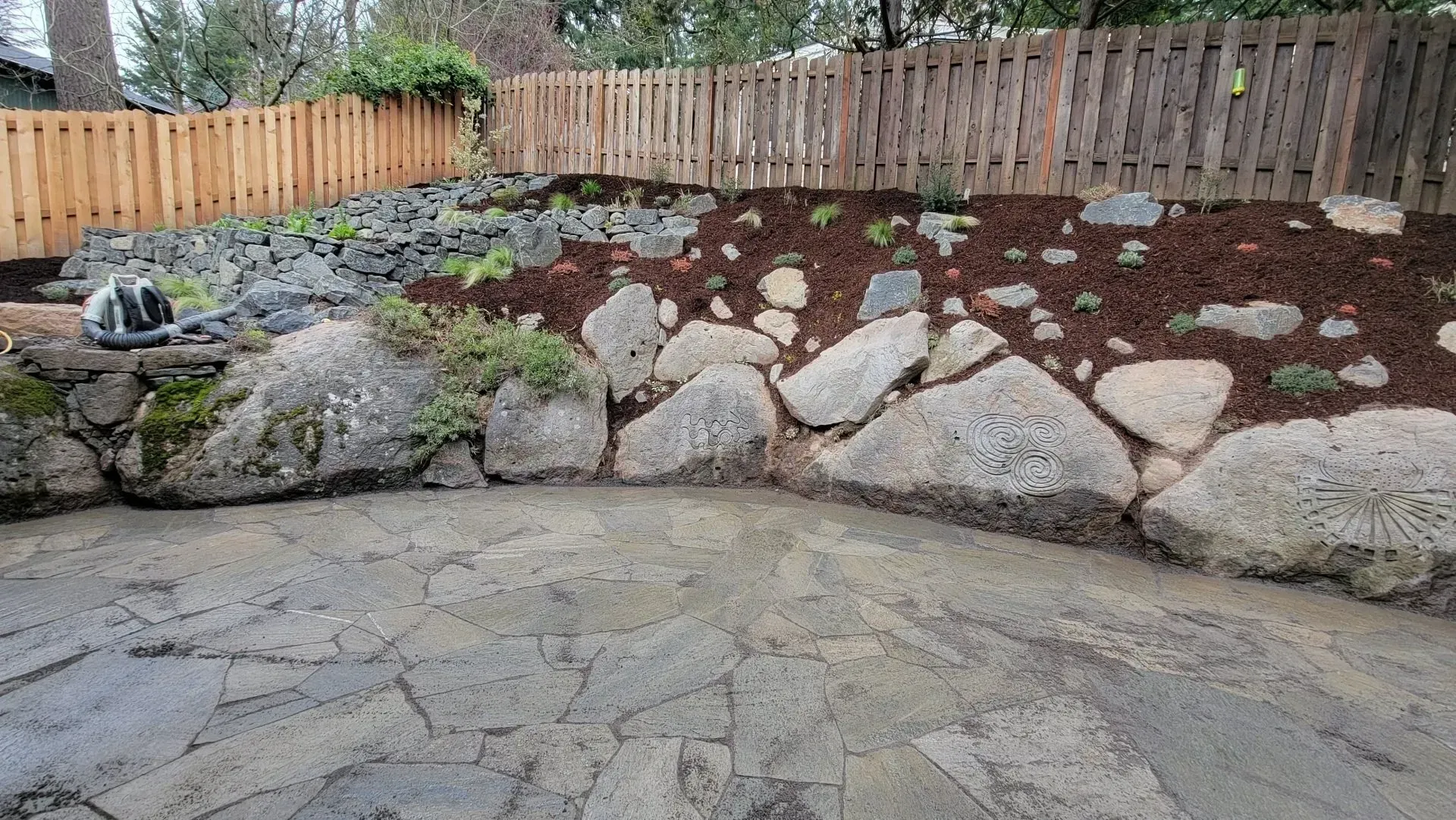 Stone patio with a rock retaining wall leading to a sloped garden bed with mulch and a wooden fence in the background.