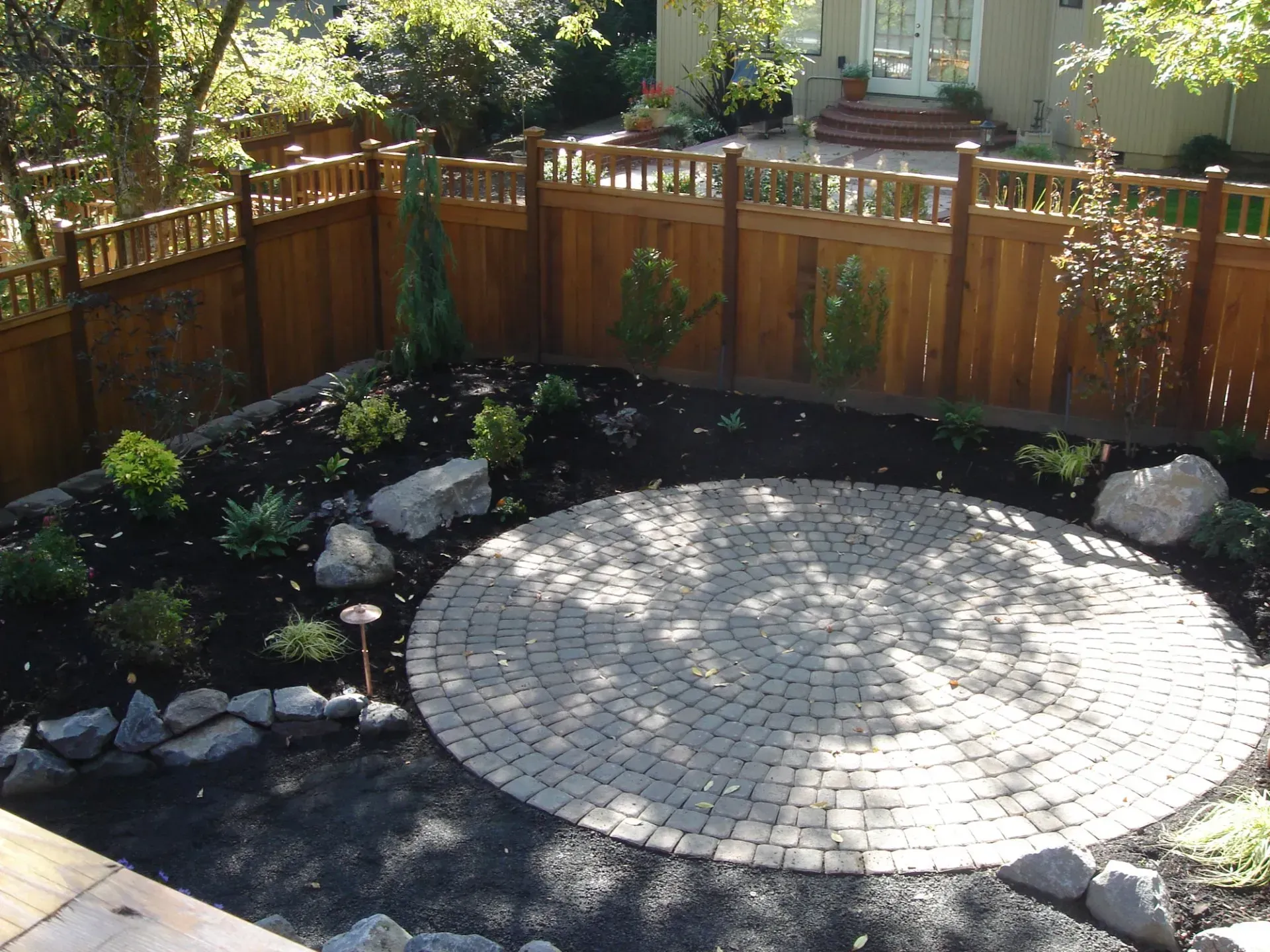 A circular brick patio surrounded by a landscaped garden with a wooden fence.