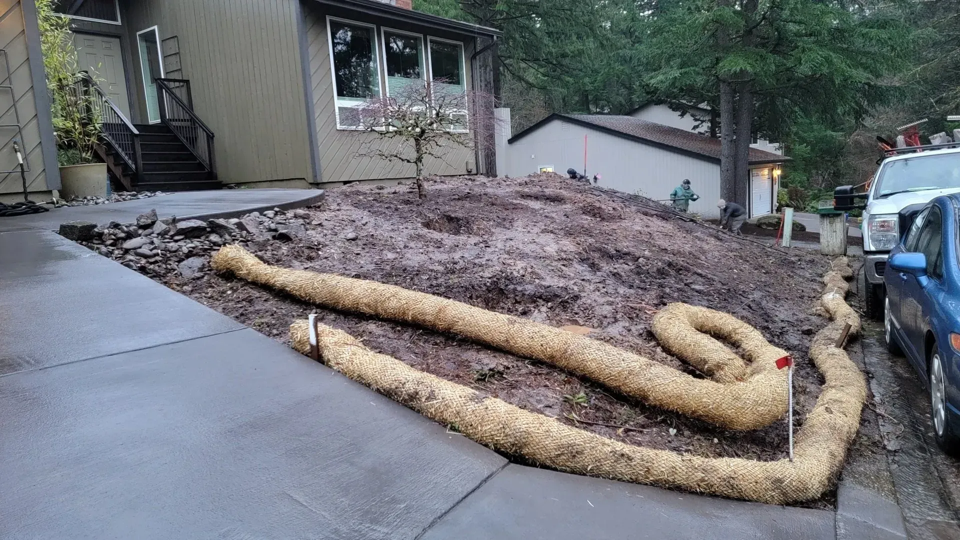 A tan-colored erosion control log is coiled around a bare hillside covered in brown mulch next to a driveway.