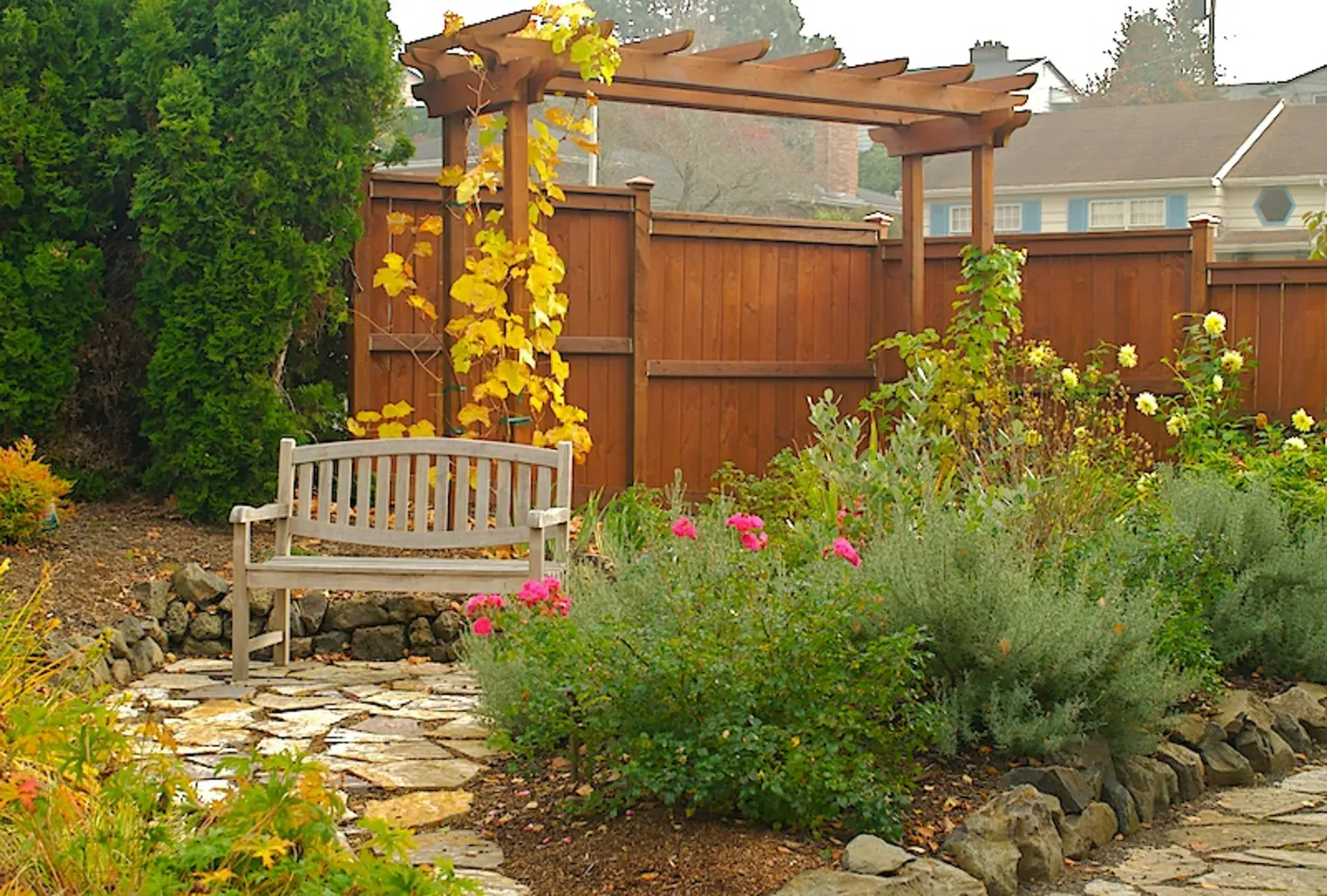 A wooden bench sits in a garden with a pergola and a wooden fence, surrounded by colorful plants.