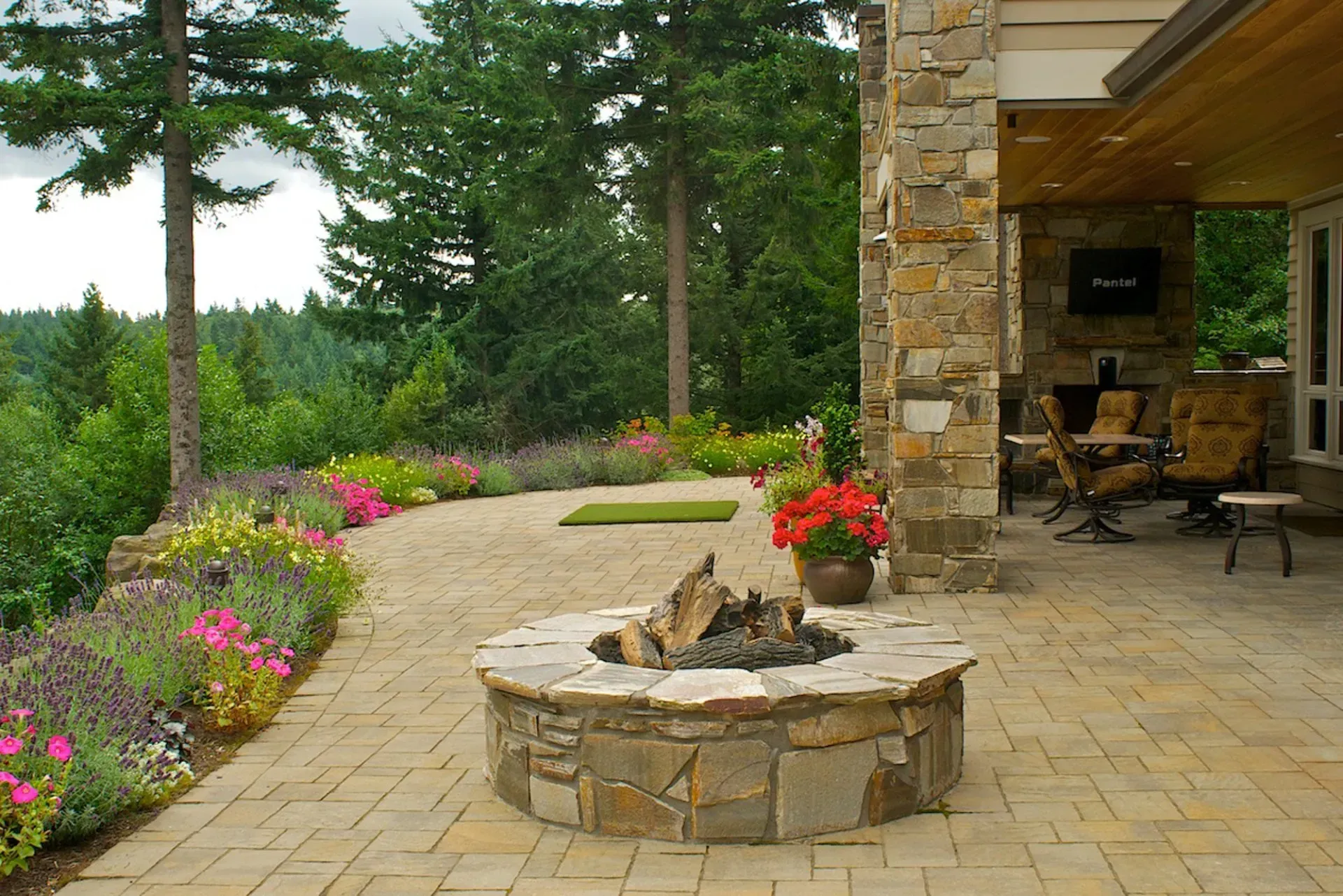 Stone fire pit on a patio with a view of a forest and blooming flowers. 
