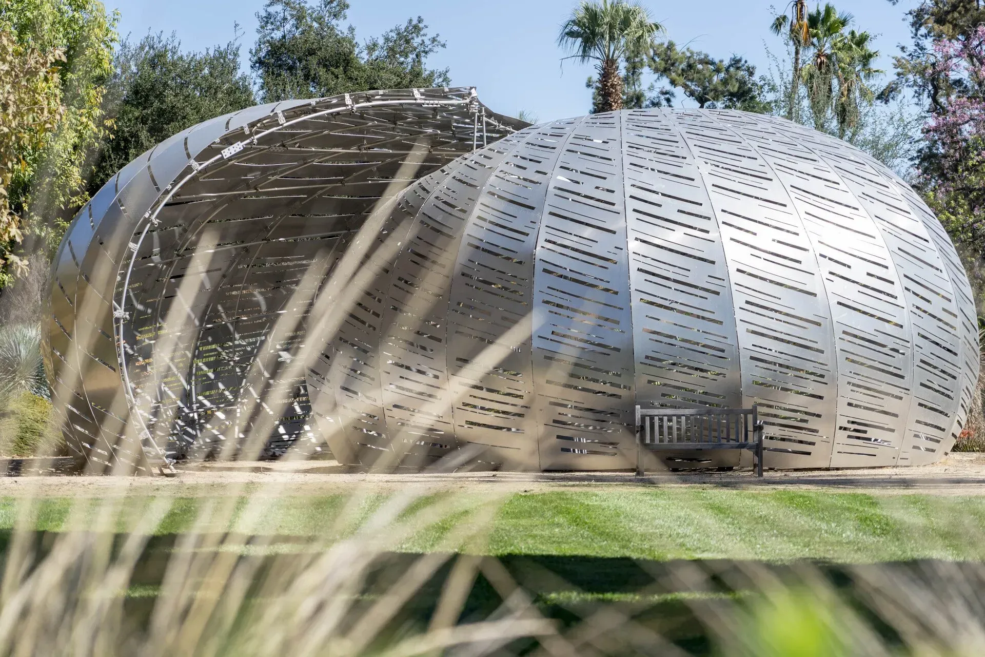 A large, modern stainless steel sculpture in a park-like setting with grassy foreground and trees.