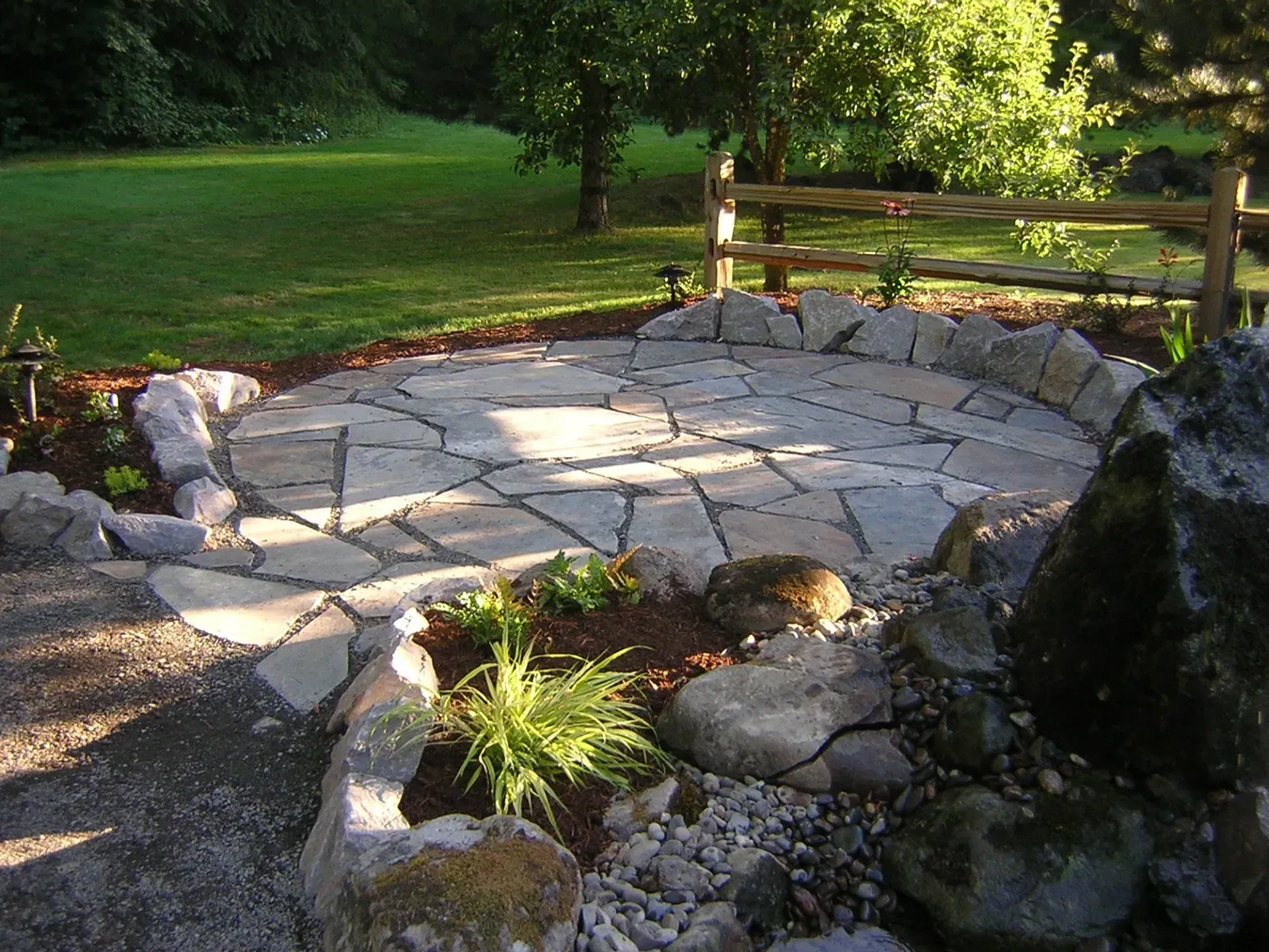 A circular stone patio in a garden, bordered by rocks and greenery, with a wooden fence and trees in the background.