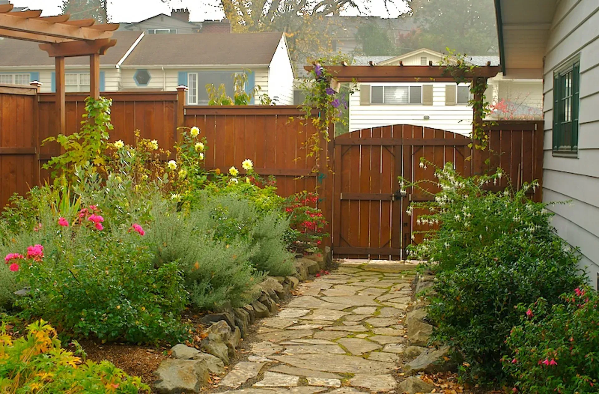 Stone path leading to a wooden gate in a garden with flowers, shrubs, and a fence. Houses are visible in the background.