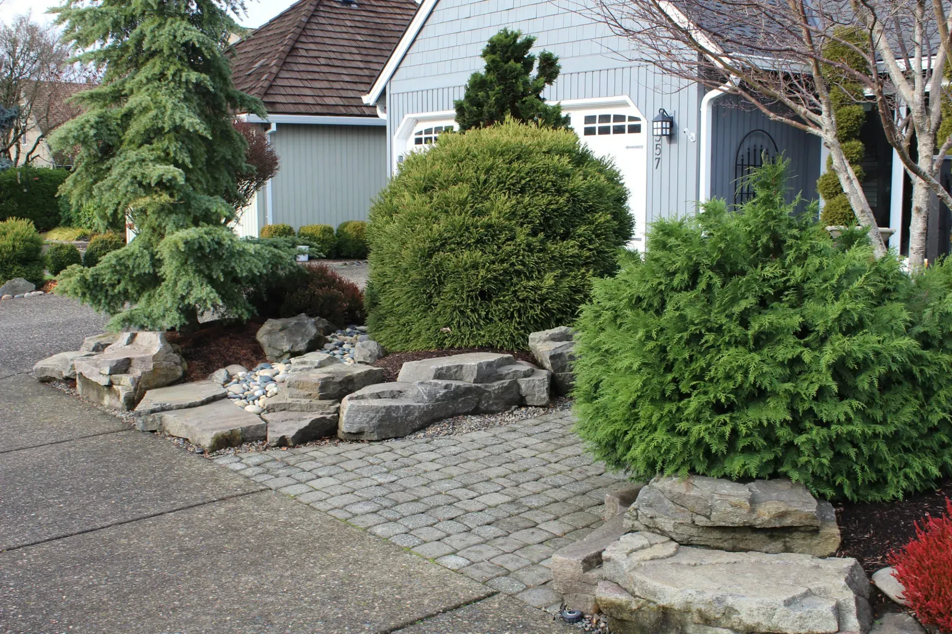 A landscaped front yard featuring a stone walkway, rock borders, and various green shrubs.